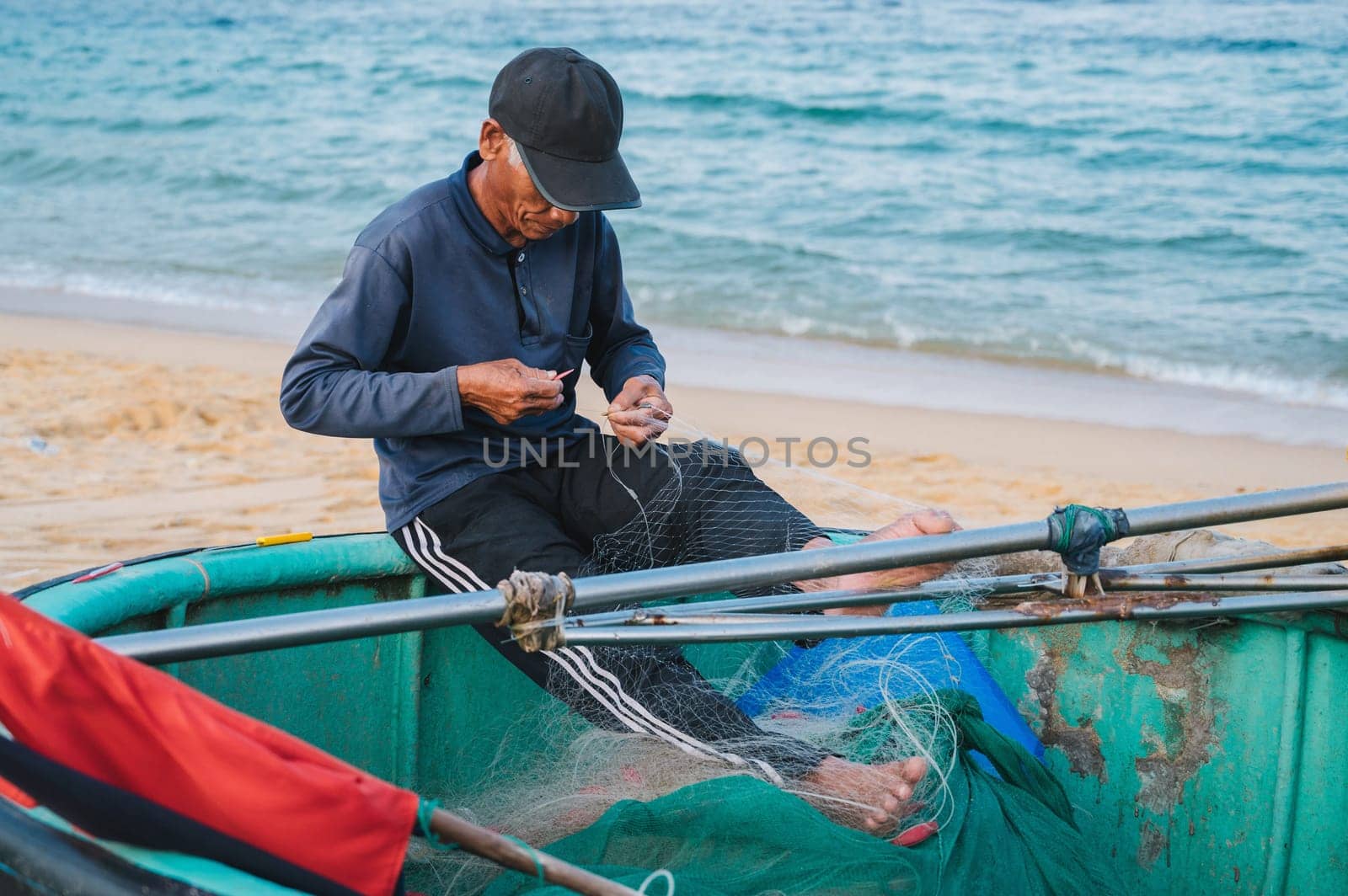 Vietnamese male fisherman repairs a fishing net sitting on a traditional basket boat on beach by sea in a fishing village in Vietnam