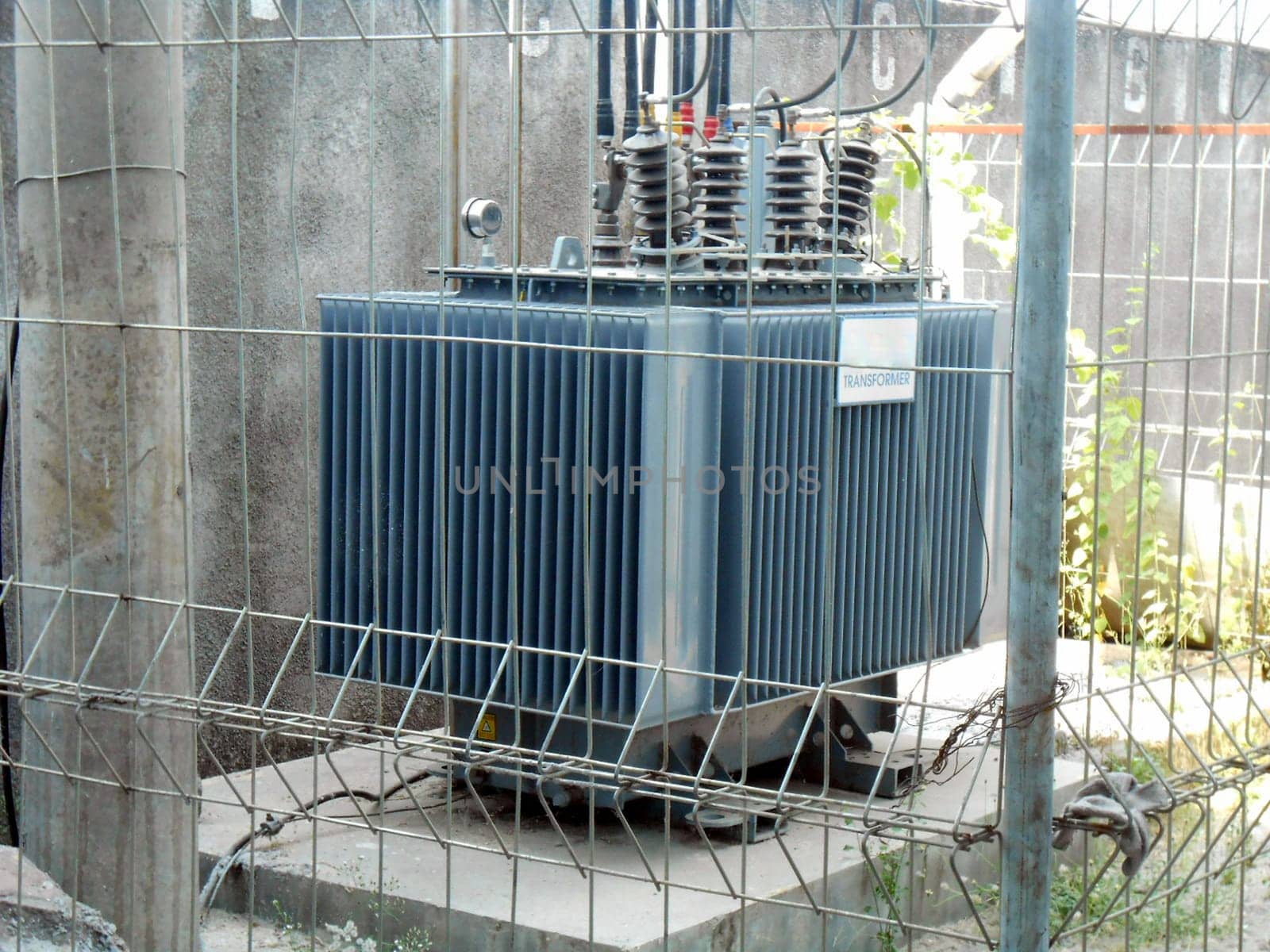 High Voltage Transformer Enclosed Behind Metal Fencing in an Industrial Setting, An image depicting a high-voltage electrical transformer protected by a secure fence in an industrial environment, emphasizing industrial infrastructure and safety measures for operational energy equipment.