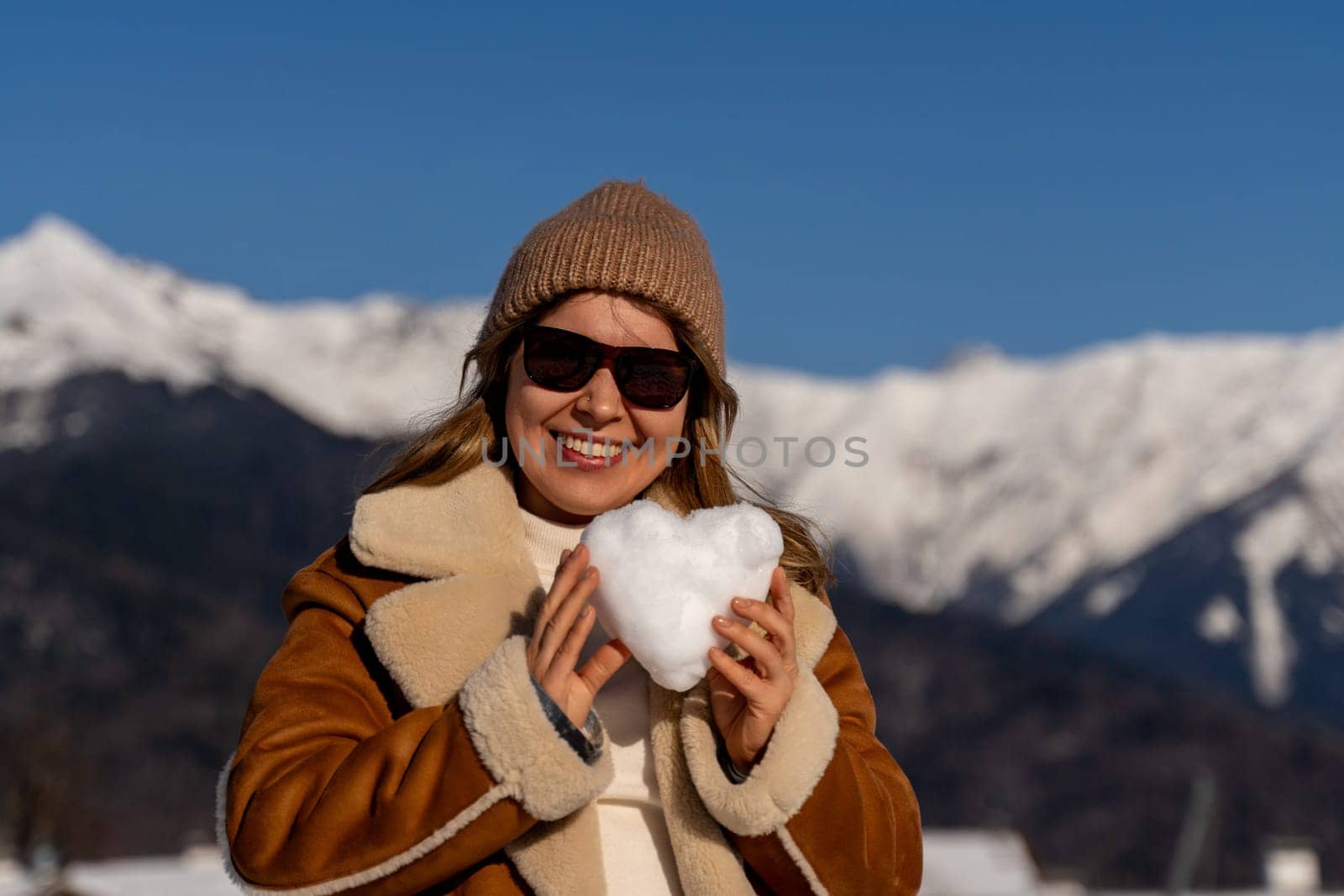 Smiling woman in sunglasses and brown coat holds a snow heart, mountains in the background. Sunny winter day.
