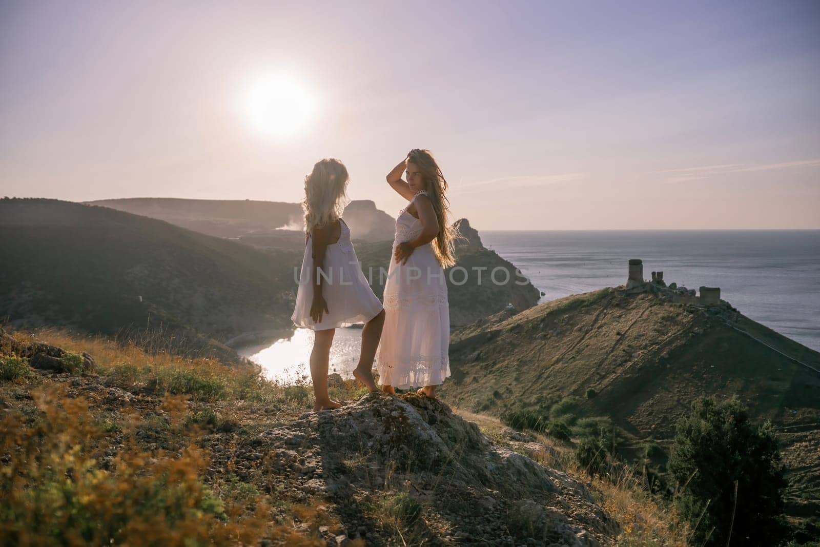 Two women are standing on a hill overlooking the ocean. They are holding hands and looking out at the water. The scene is peaceful and serene, with the sun shining brightly in the background