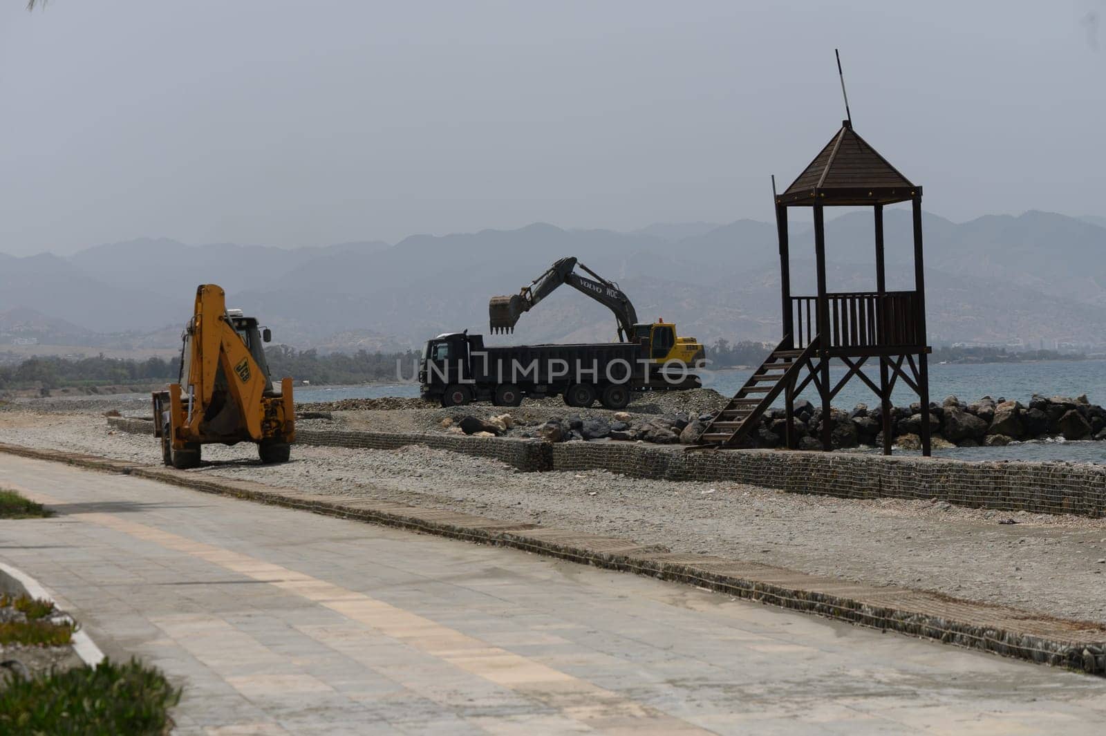 Heavy-duty yellow excavator loads large sea rocks into a dump truck on the Mediterranean coast of Cyprus during shoreline construction.