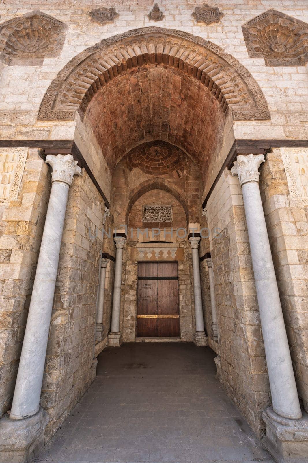 Exterior view of the large arched entrance to the Mosque of al-Zahir Baybars in Cairo, Egypt. Architectural details such as the stonework, arches, and columns are visible