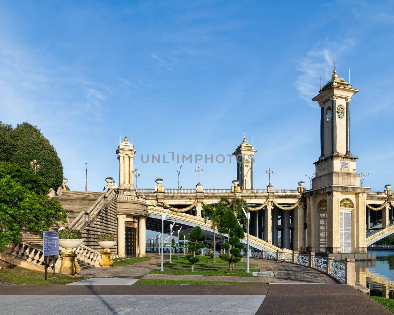 The Seri Gemilang Bridge, an architectural landmark in Putrajaya, Malaysia, showcases ornate towers, arches, and a landscaped park by Putrajaya Lake under a sunny blue sky