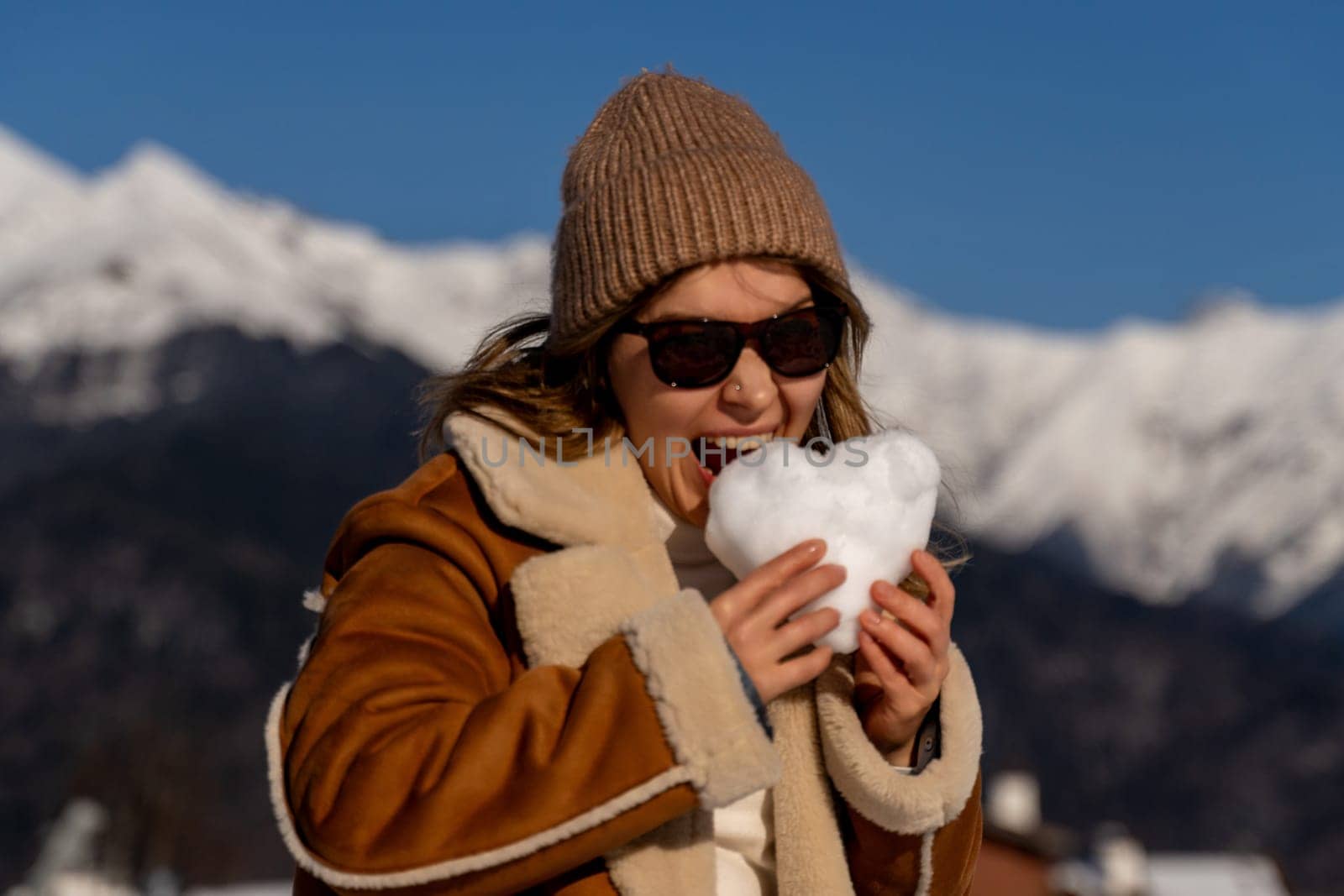 Woman in brown coat and knit hat playfully bites a heart-shaped snowball. Mountains form a scenic backdrop on a sunny day.