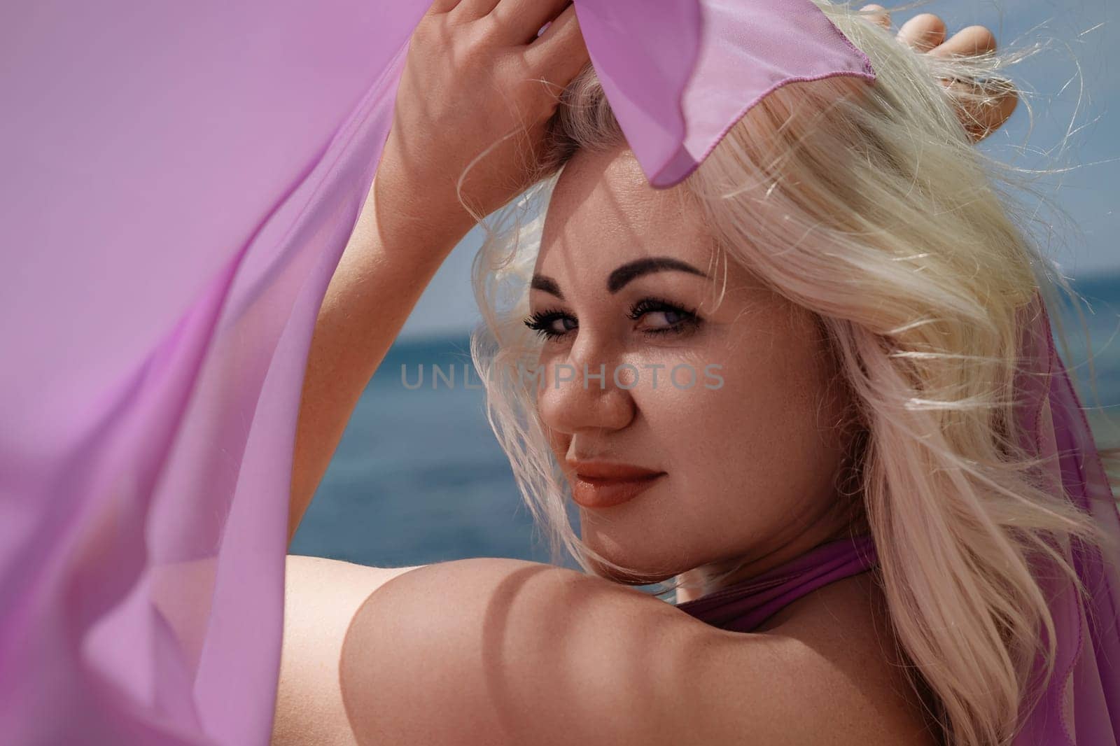 A blonde woman poses outdoors with a sheer purple fabric. Ocean view in the background.