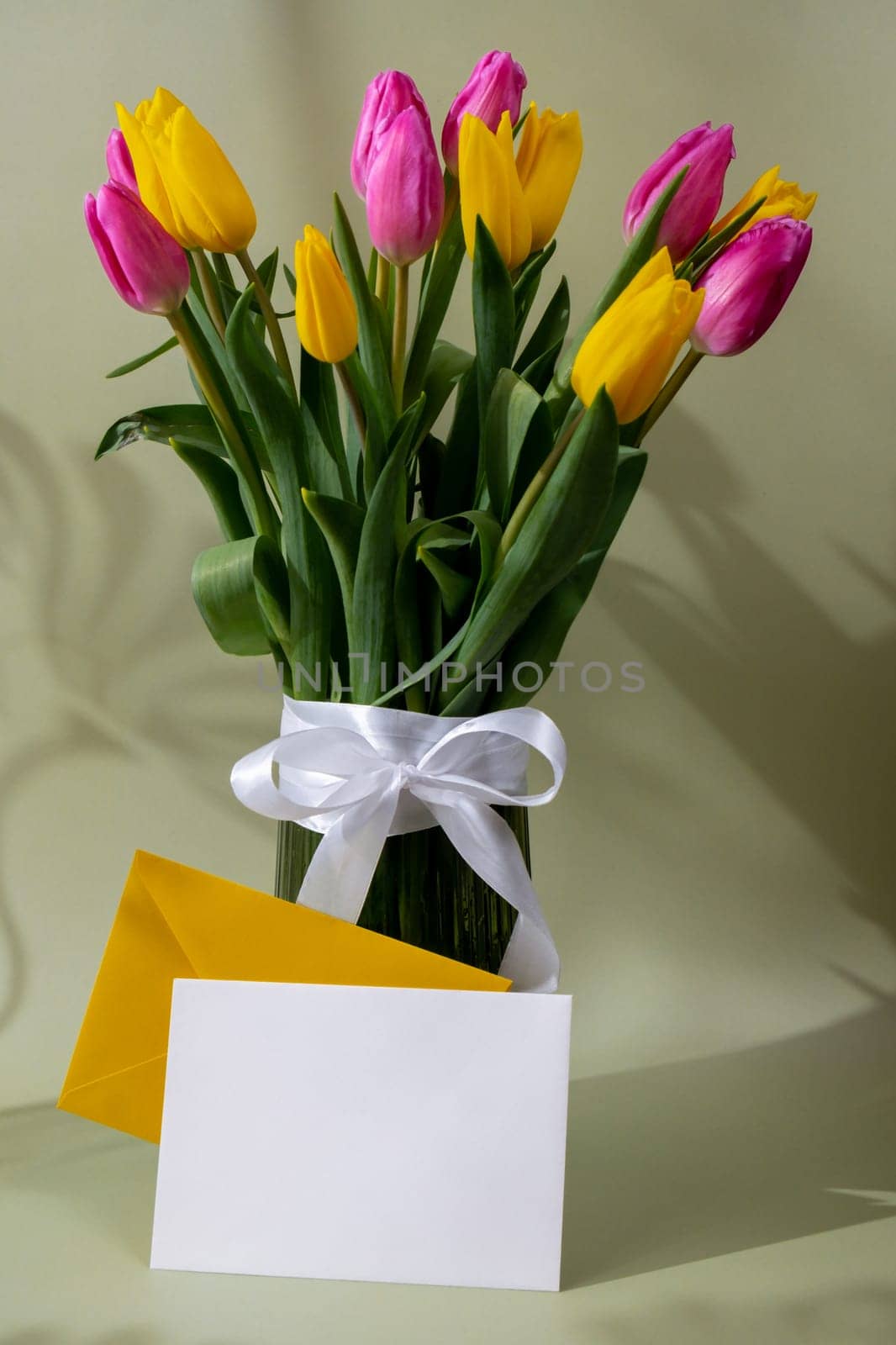 Empty white envelope mock up template Bouquet of colorful tulips in vase in pastel green background. Copy space for greeting card. Holiday flowers springtime atmosphere