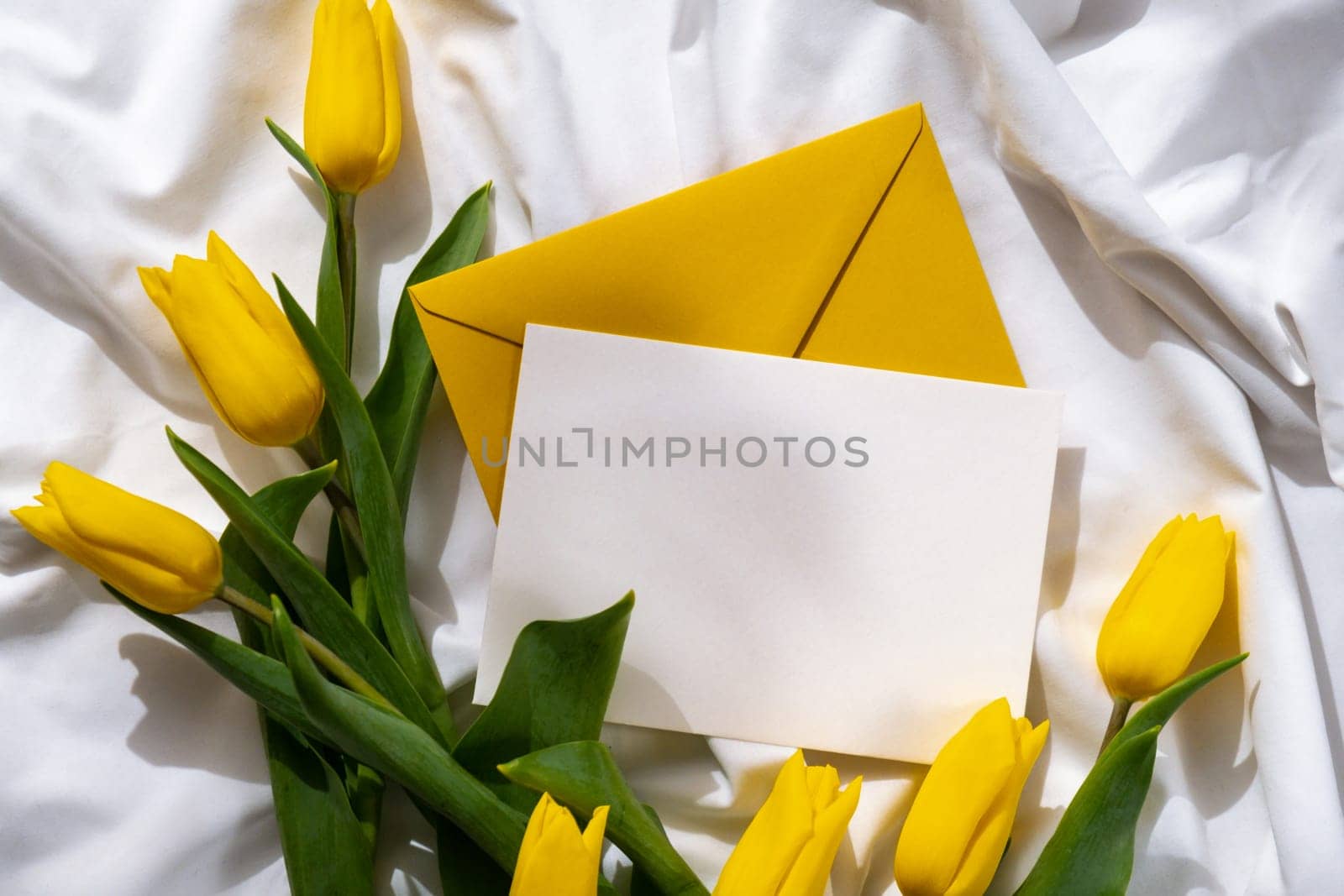 Mock up of white empty envelope greeting card with yellow fresh tulips on white bed with trendy deep shadows. Spring flowers in yellow colors. Romantic postcard wallpaper design Copy space
