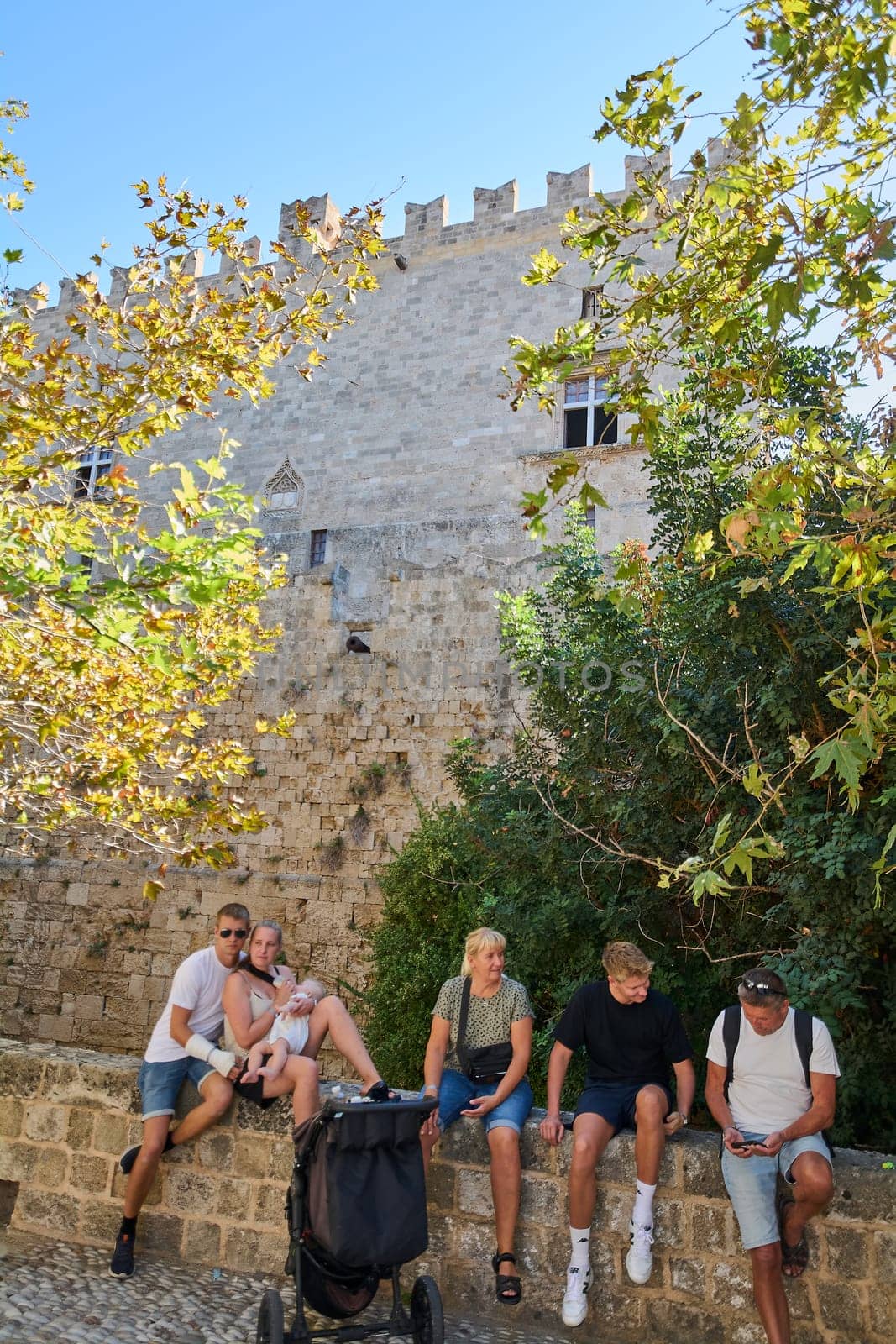 Rodas.Greece - june 01, 2025: A group of friends and a family with a baby relax on a low stone wall enjoying the sunny weather in front of the formidable medieval Grossmeisterpalast, partially obscure