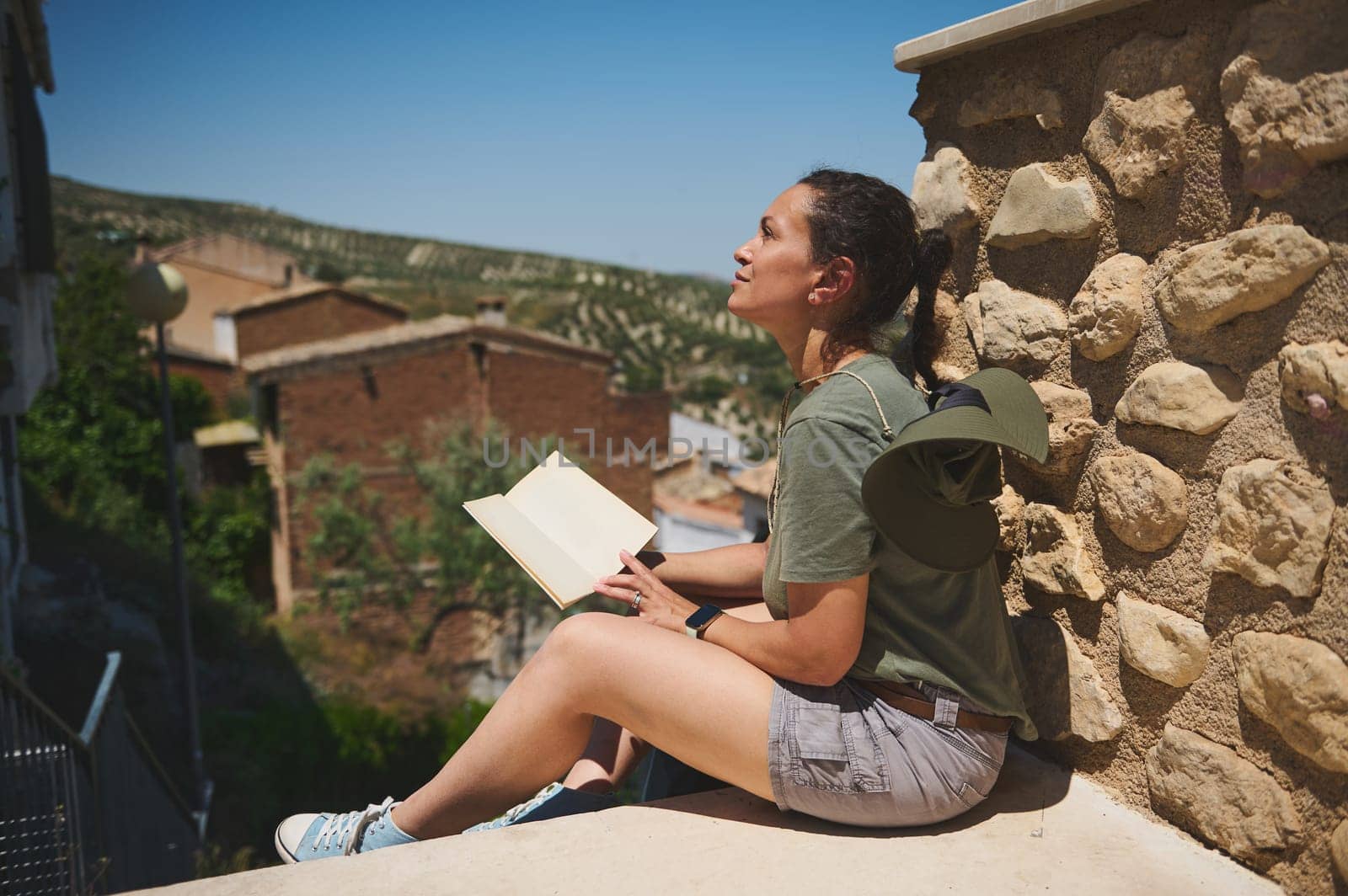 A traveler enjoying the outdoors while resting against a stone wall with an open notebook. The setting features a rustic village and picturesque greenery, embodying a peaceful and contemplative mood.