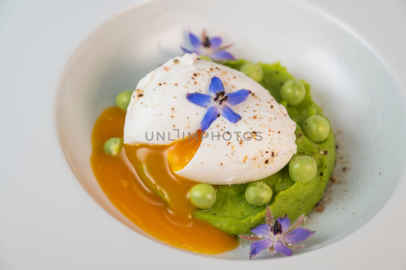 A fresh pea puree is beautifully plated, featuring a poached egg at the center and adorned with borage flowers. The dish showcases vibrant colors and textures, perfect for any spring menu.