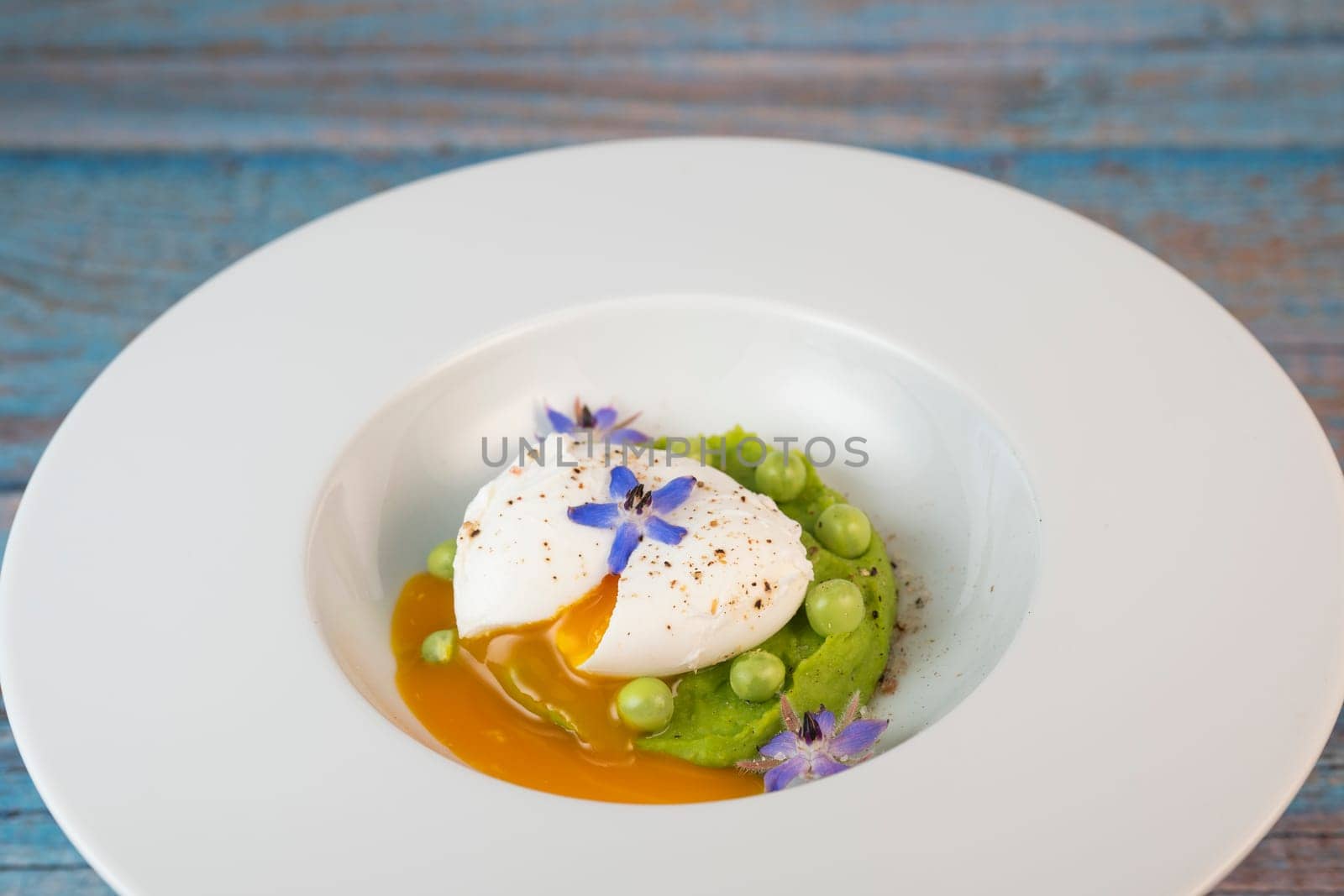 A fresh pea puree is beautifully plated, featuring a poached egg at the center and adorned with borage flowers. The dish showcases vibrant colors and textures, perfect for any spring menu.
