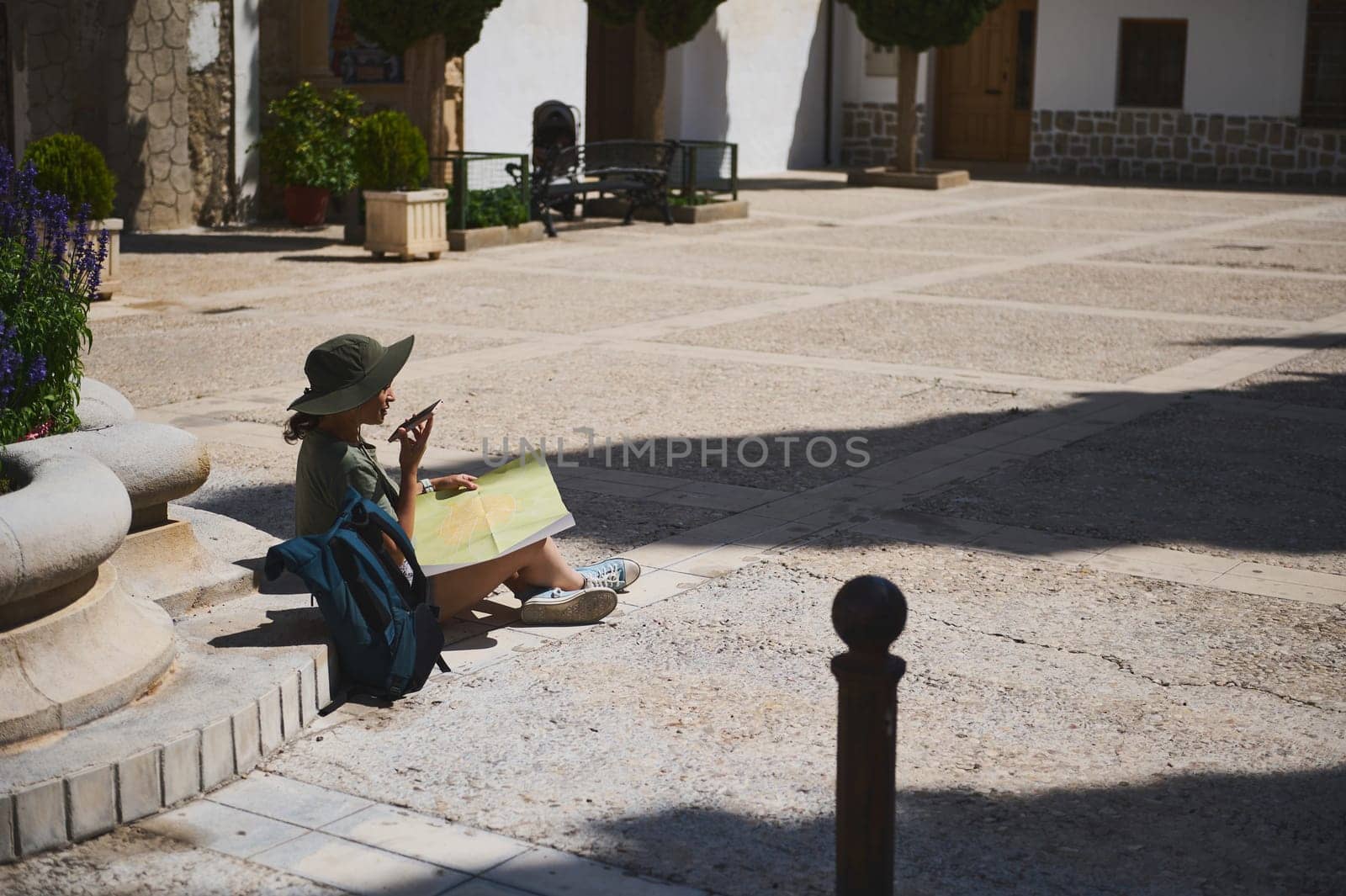 A lone traveler sitting in a historic plaza, examining a map under the bright sunlight, exudes an essence of adventure and discovery in a scenic and tranquil town.