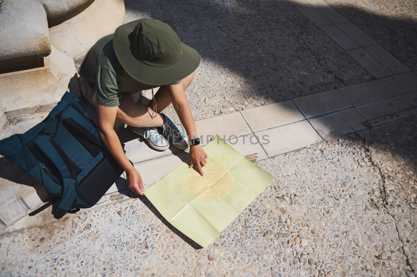 A backpacker is sitting on a sunny tile street, studying a city map, planning their travel route through a charming historic town with medieval architecture.