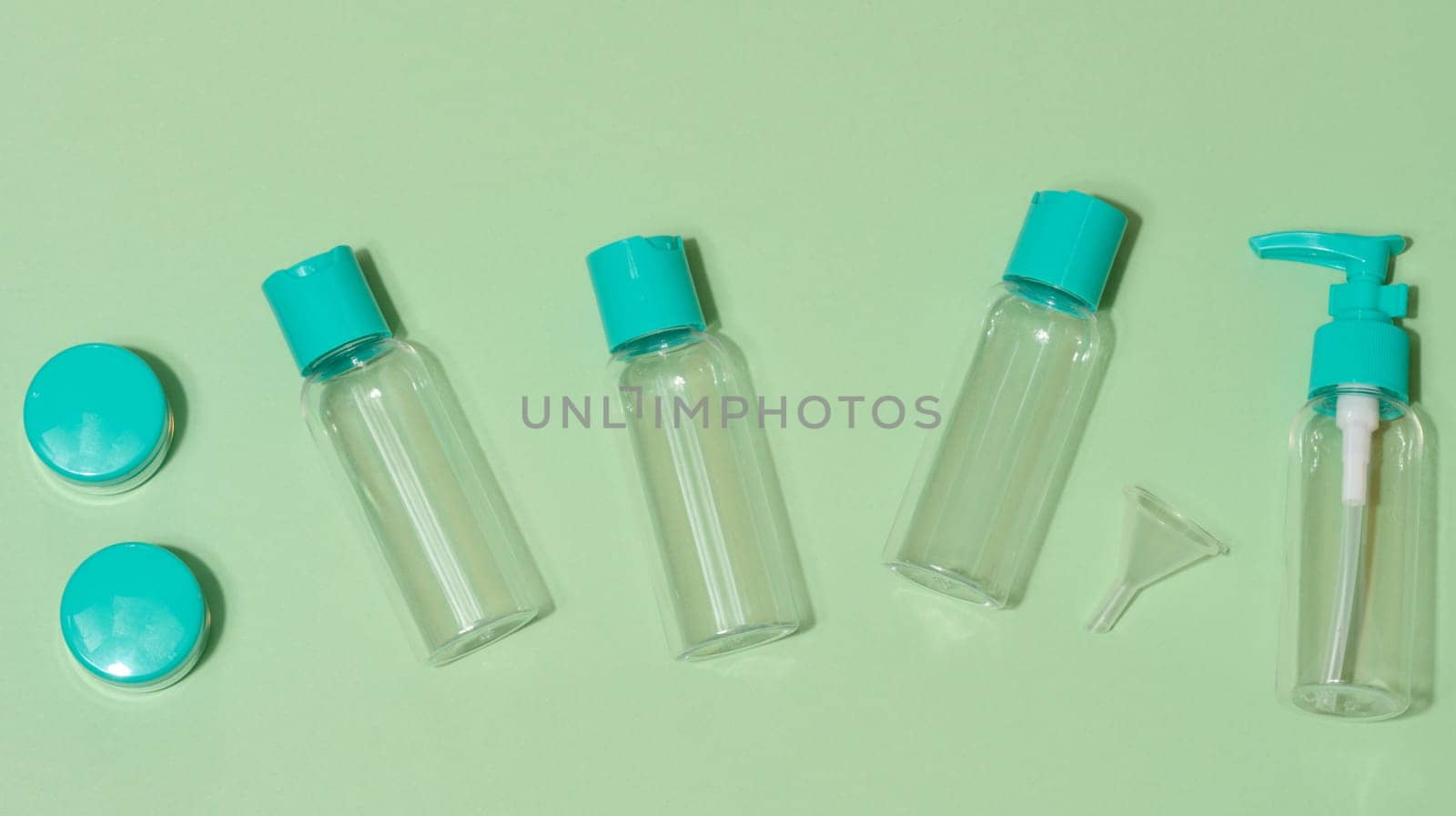Top view of set of five transparent cosmetic bottles and two round jars with turquoise lids and one funnel, light green background