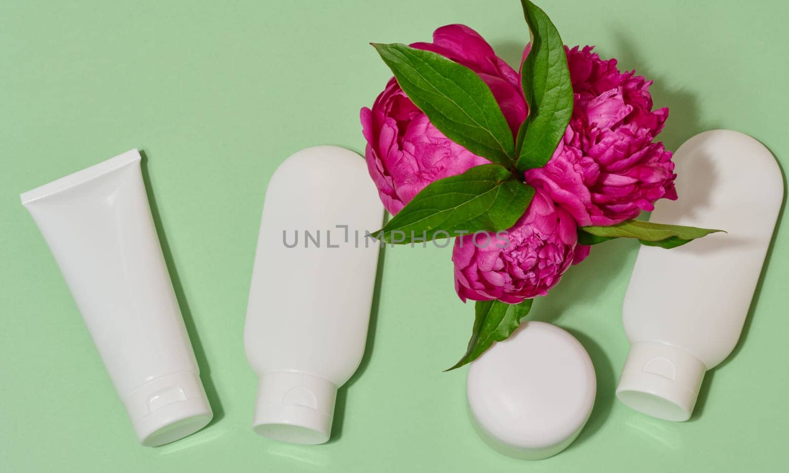 Top view of cosmetic tubes without labels in white packaging and pink peonies with green leaves, light green background