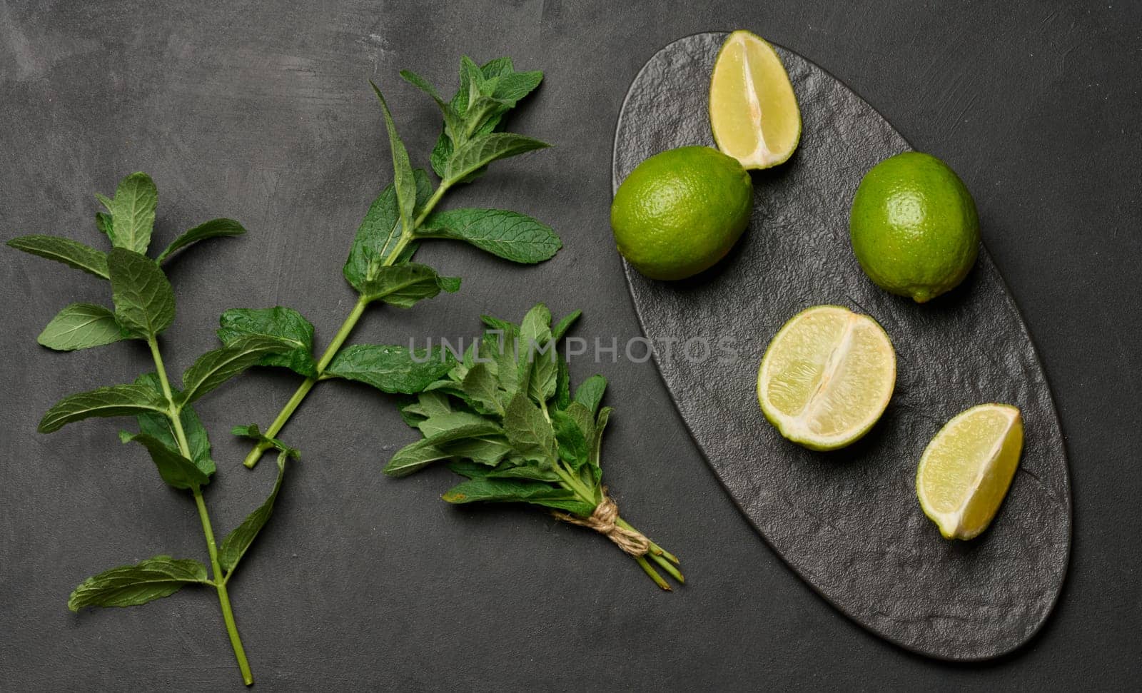Whole and cut limes with fresh mint leaves arranged on a dark textured surface. Composition perfect for food, drink, or summer themes
