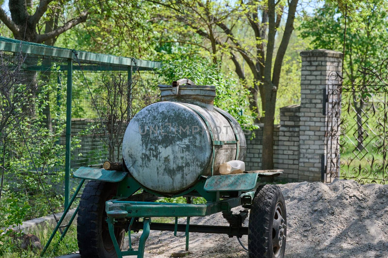 An old metal tank on a trailer, covered with traces of rust and time, stands against the background of green trees and a chain-link fence. The trailer is painted a worn green color