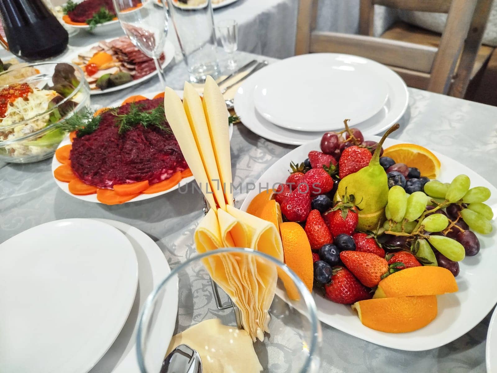 Various dishes are displayed on a table set for a celebration. Several white plates, salads, sliced meats, and a fruit platter with grapes, strawberries, oranges, and a pear are present.