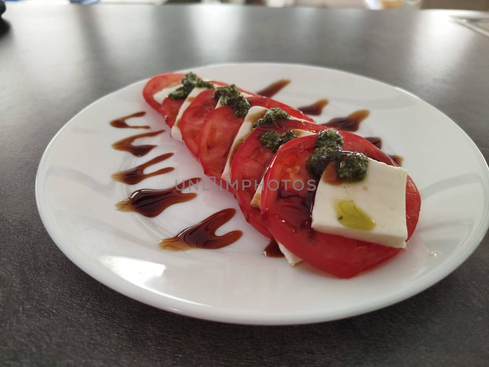 A Caprese salad is presented on a white plate. Red tomato slices alternate with white cheese, decorated with pesto and balsamic glaze.