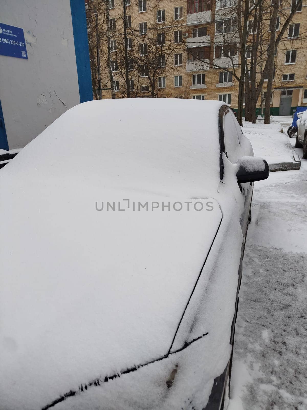 Car Covered in Snow after Snowfall. High quality photo A grey car is completely covered in fresh white snow, parked outside a residential building during a winter day. Trees without leaves and more snow visible in the background.