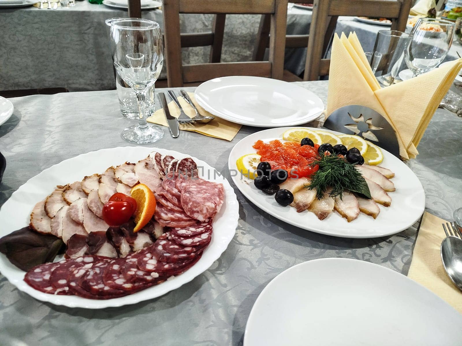 Two platters, one with assorted meats and the other with fish, lemons, olives, and dill, are placed on a set banquet table for guests to enjoy at an event.