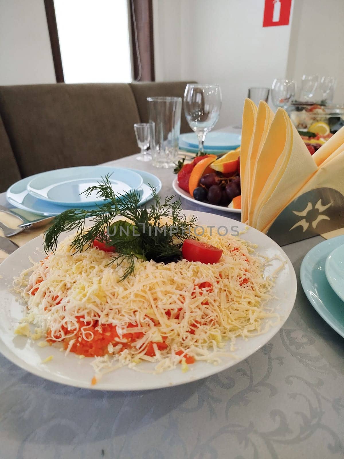 A table setting is seen with salad, fruit, plates, glasses, and folded napkins. The layered salad is topped with grated cheese, tomato pieces, and dill. This dining location may be in Russia.