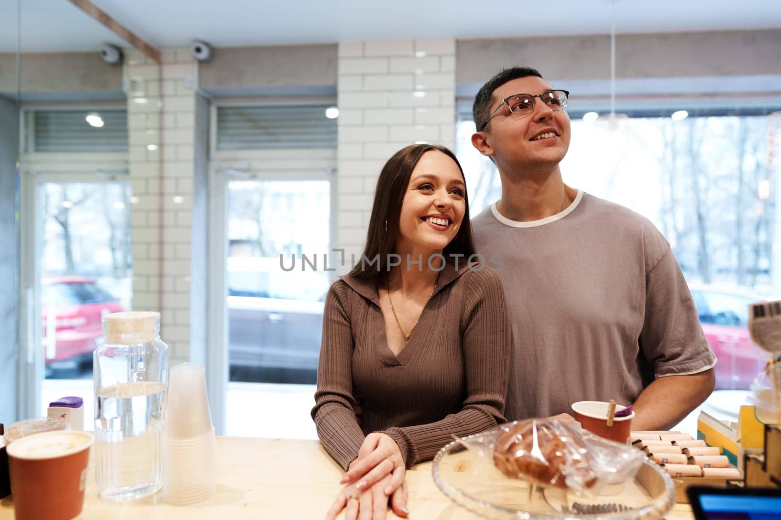 A couple stands together at a cafe counter, smiling and looking out the window. They are surrounded by coffee cups and baked goods, creating a warm and inviting atmosphere.