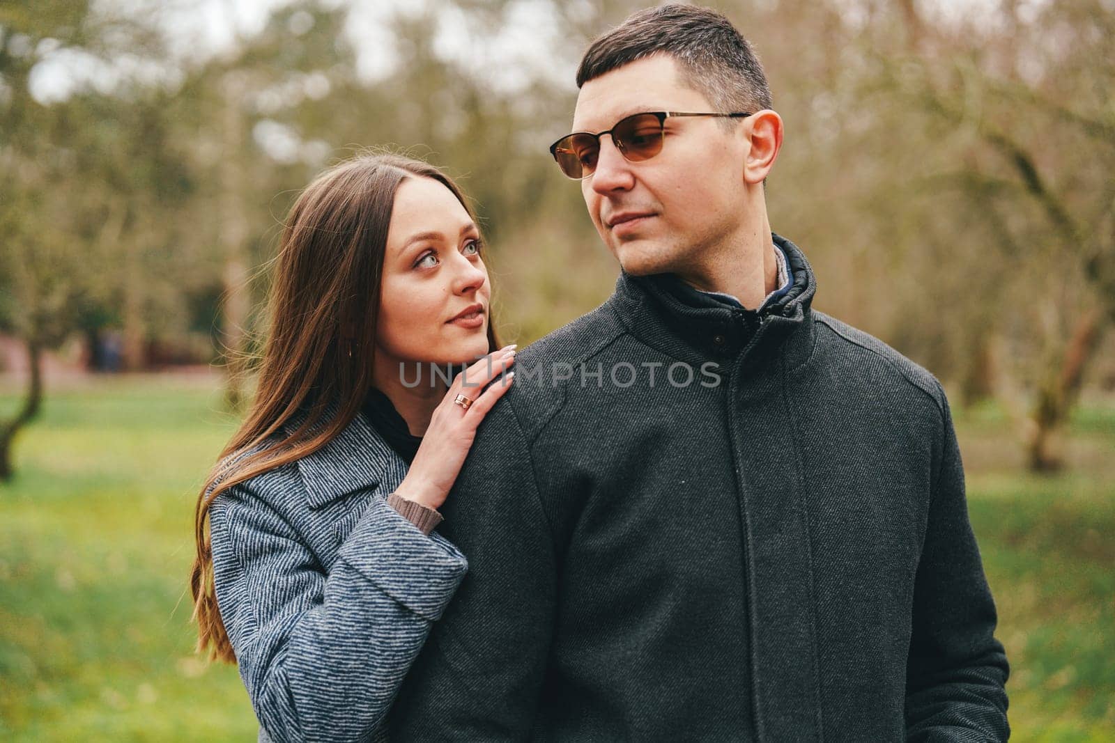 A couple stands close to each other in a lush green park. The woman gazes affectionately at the man, who is wearing sunglasses and a stylish coat.