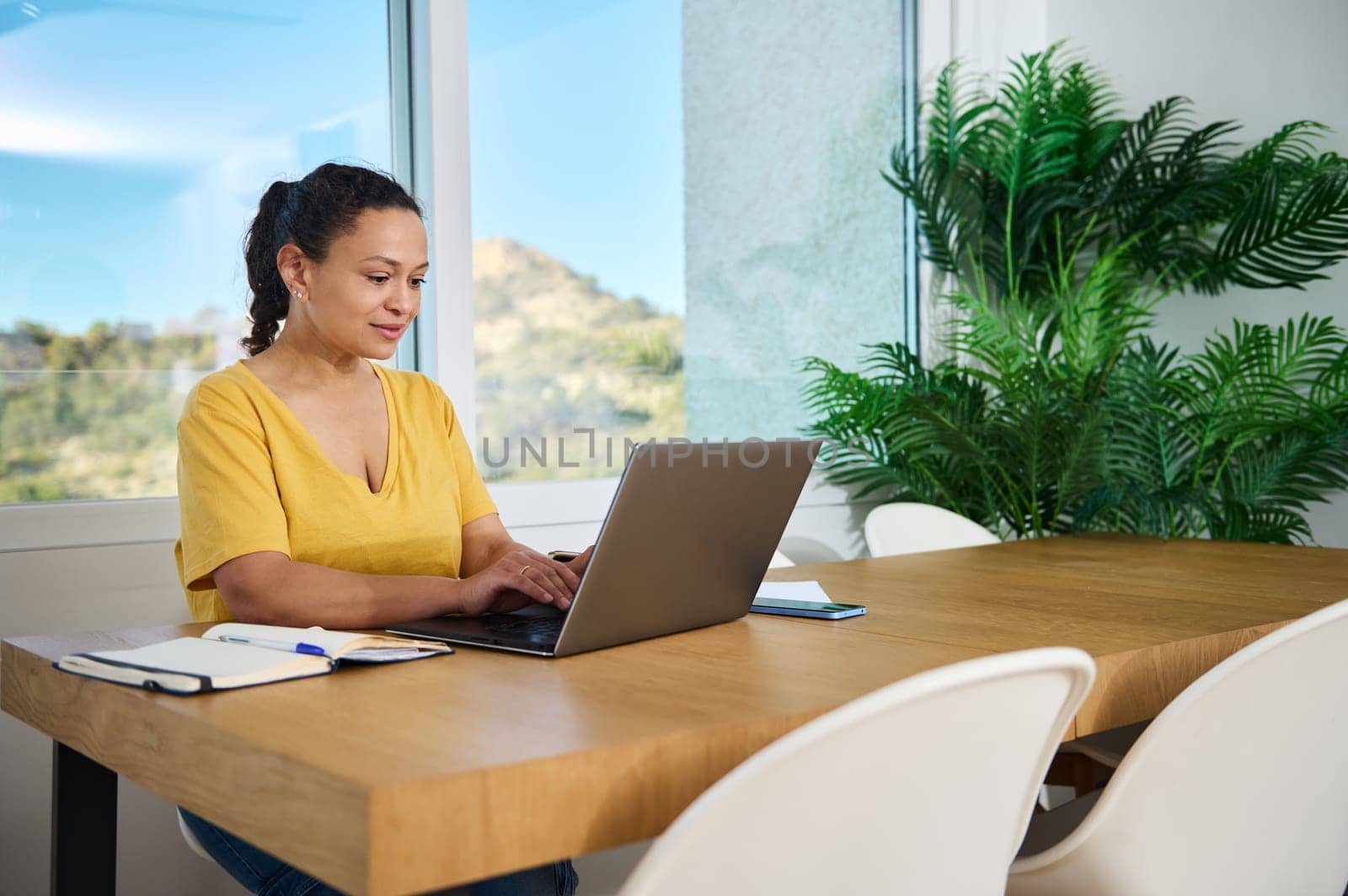 A focused woman working on her laptop in a modern, well-lit home office environment, showcasing a balance of productivity and comfort.