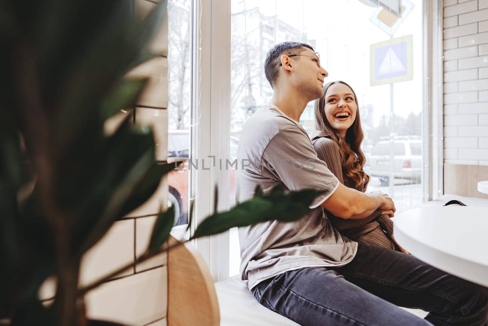 A couple sits closely at a table in a warm cafe, sharing smiles and laughter. Soft light filters through the windows, creating a relaxed atmosphere on a chilly day.