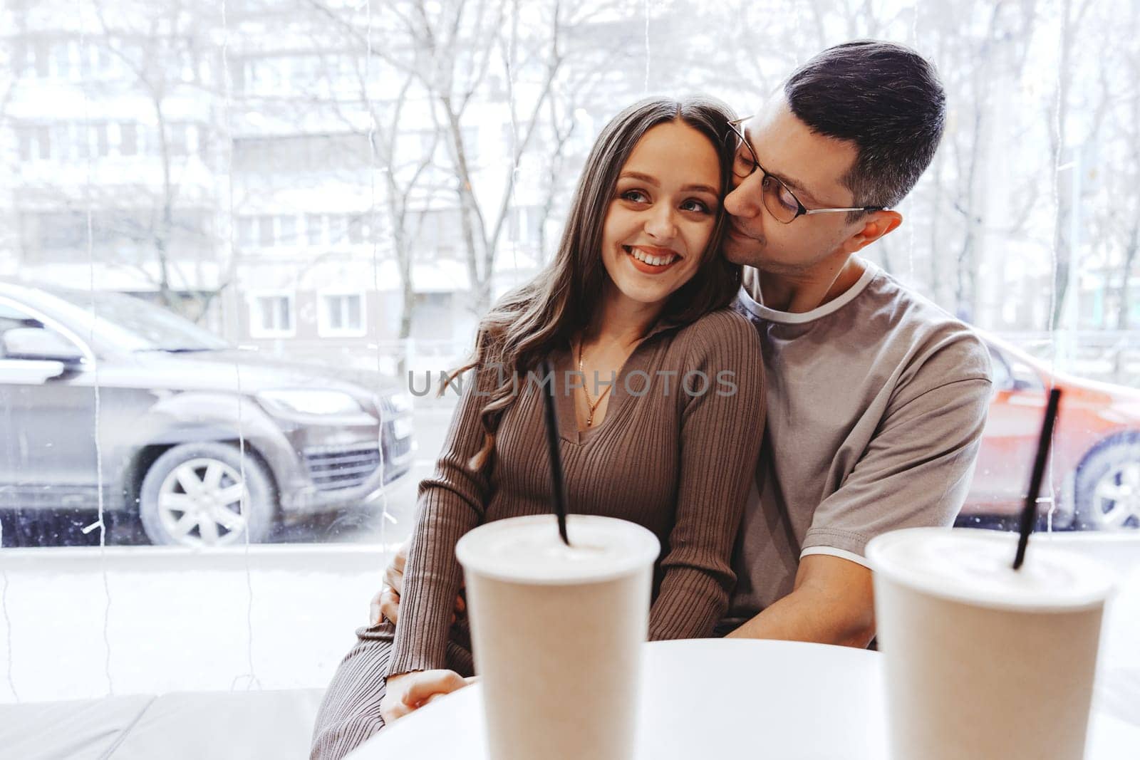 A young couple shares an affectionate moment at a cafe, surrounded by a winter scene. They sit closely together, smiling and sipping their drinks, creating a warm atmosphere.