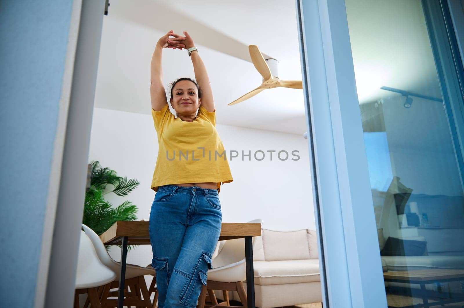 A cheerful woman stretches joyfully in her modern home interior, balancing work and leisure in her comfortable space, filled with natural accents and stylish decor.