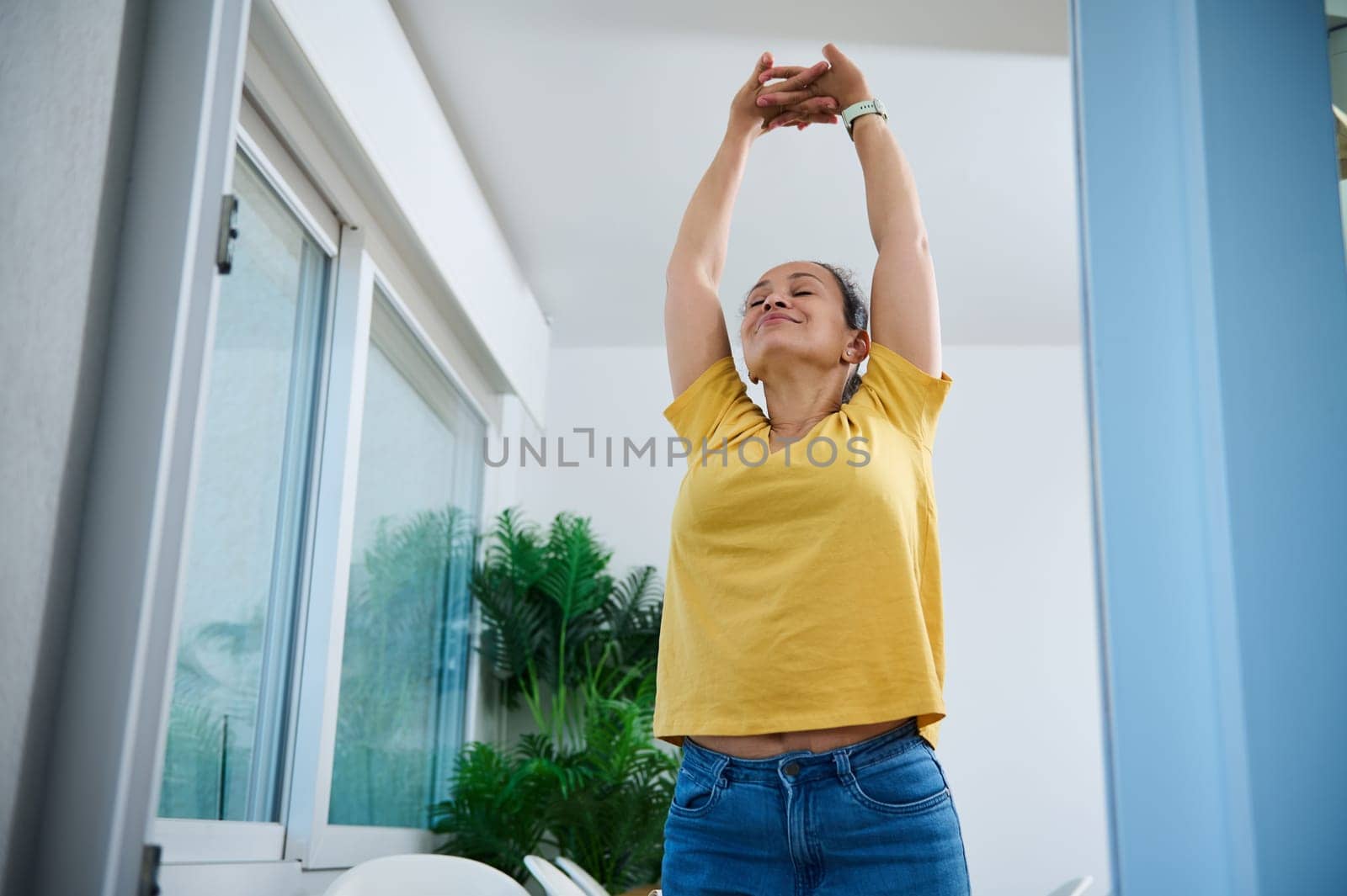 Woman in casual attire stretches inside a cozy, well-lit home space. Green plants and large windows create an environment of relaxation and vitality.