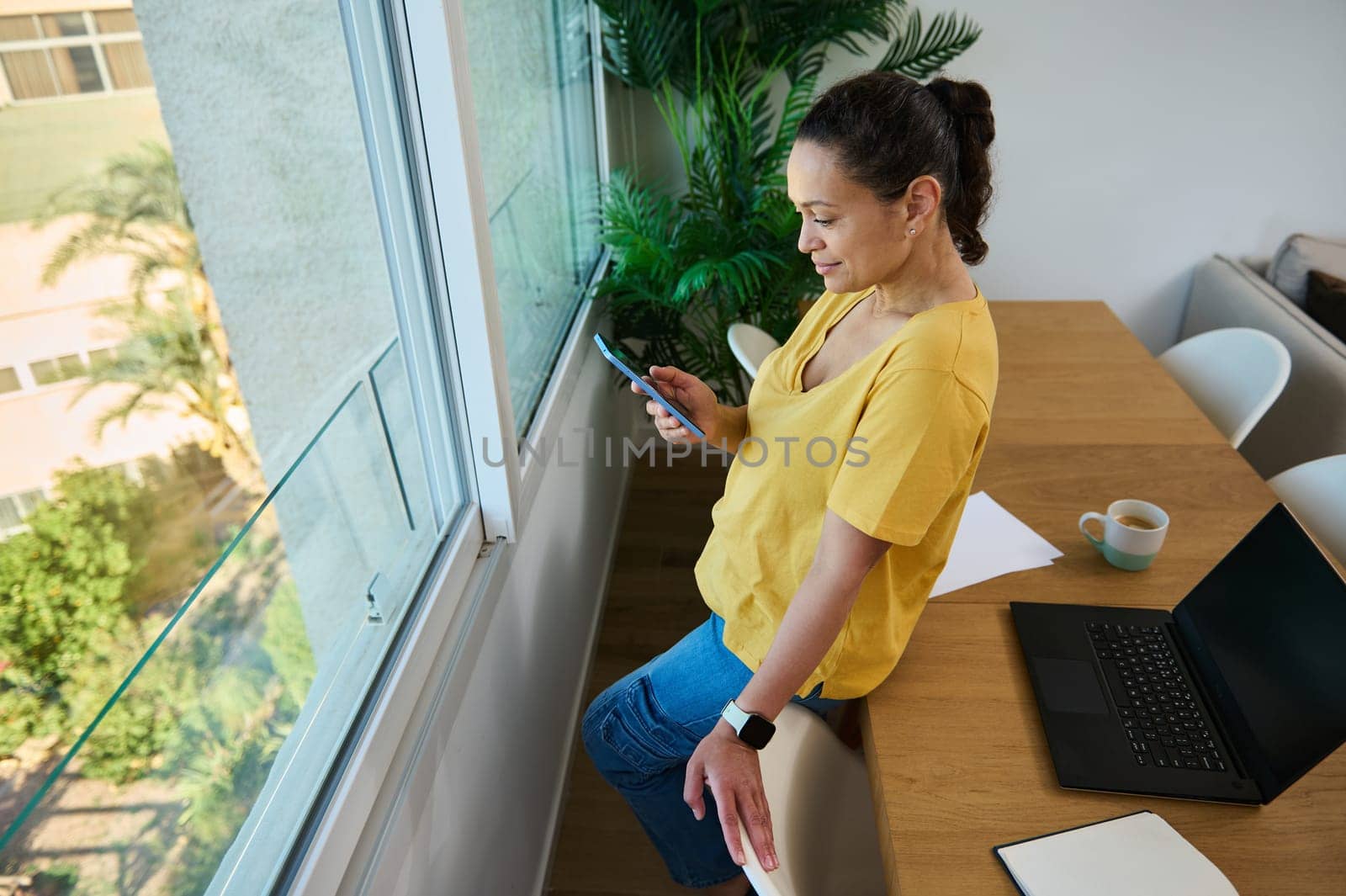 A woman leans by a window, using a smartphone during a break from work. She is in her home office setting with a laptop and coffee on the table nearby.