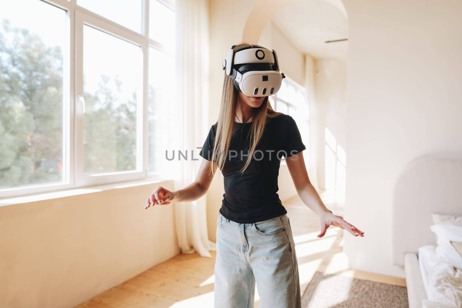 A young woman wearing a virtual reality headset stands in a spacious, sunlit room. She appears engaged and curious, moving her arms while exploring a digital world.
