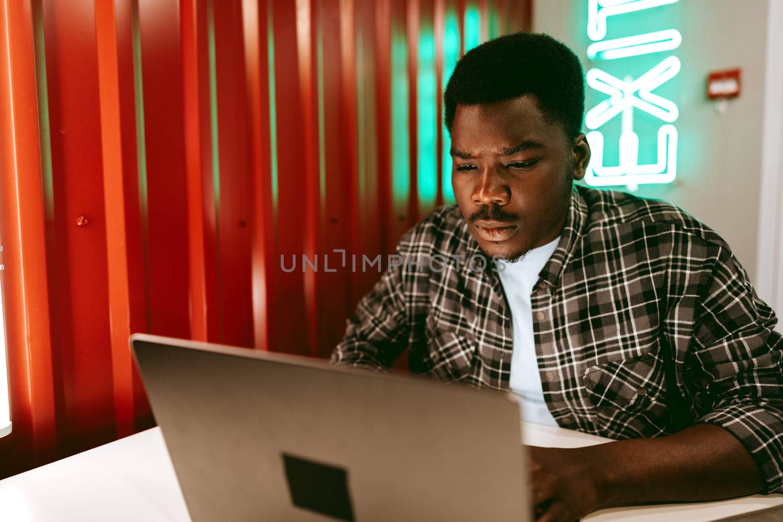 A young man sits at a table in a contemporary cafe, concentrating on his laptop screen. Neon lights illuminate the background as he engages in his work.
