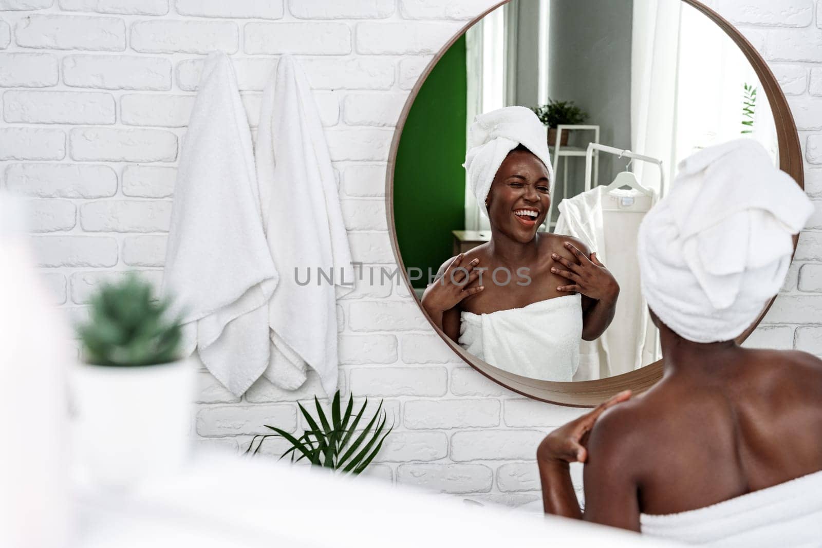 A young woman smiles at her reflection in a circular mirror while wrapped in a towel. The modern bathroom features white walls and greenery, creating a serene atmosphere.