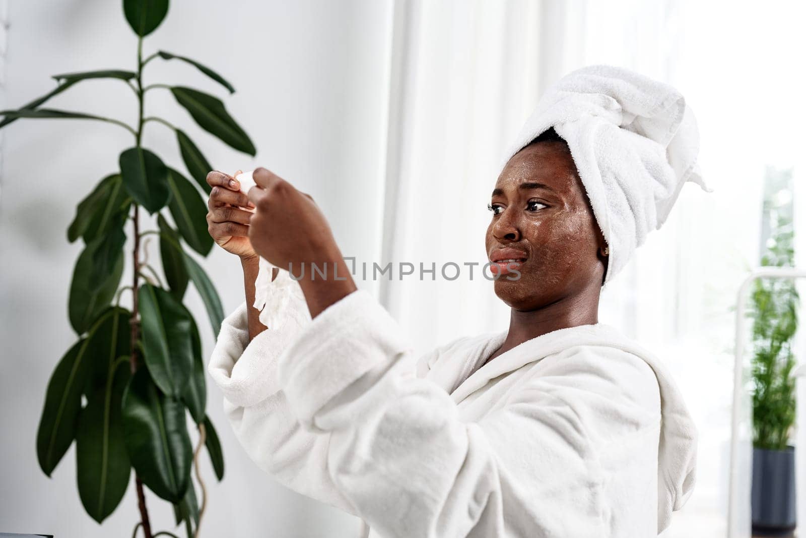 A woman wearing a white towel is applying a skincare treatment while looking in the mirror. The bathroom has bright lighting and green indoor plants, creating a refreshing atmosphere.