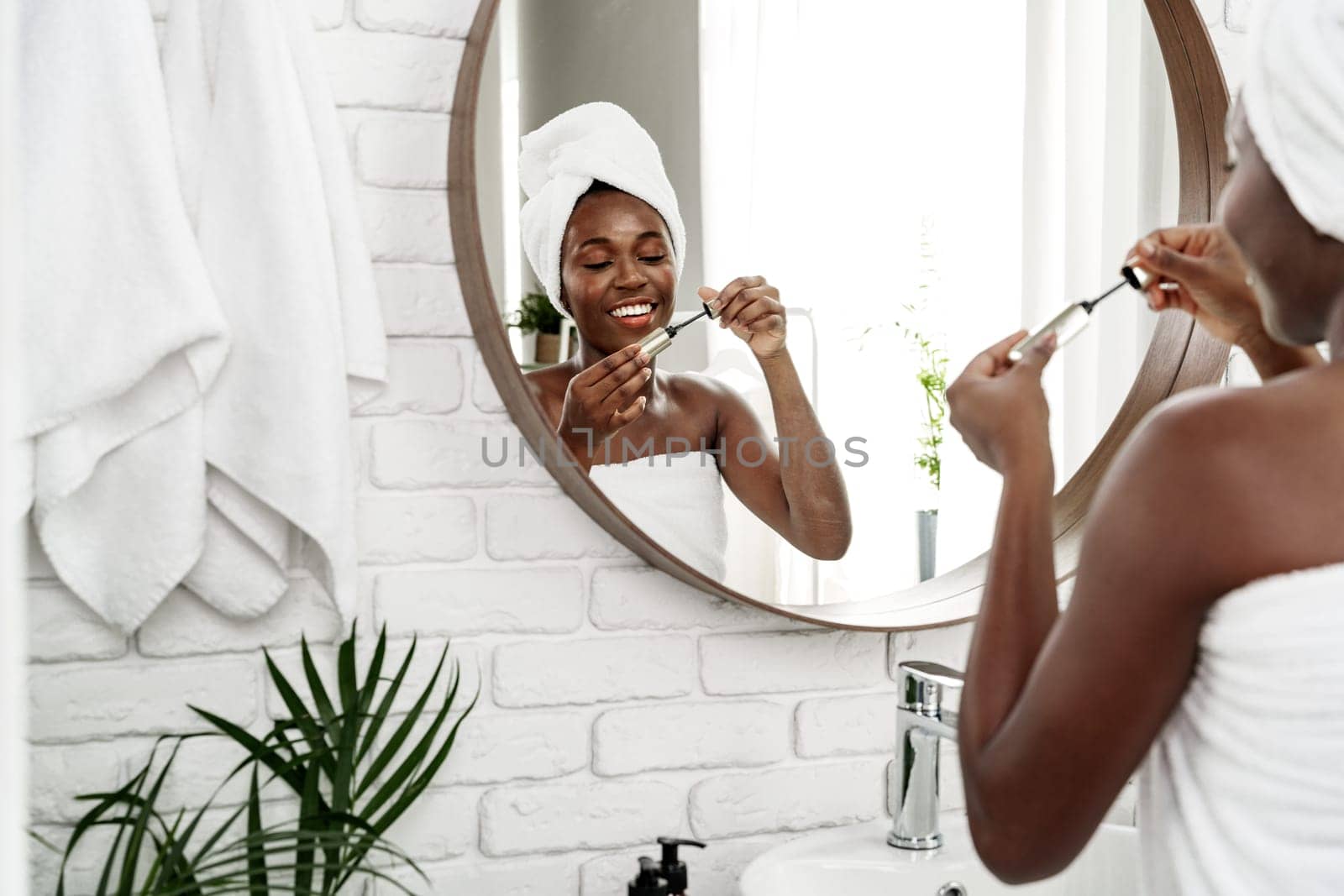 A woman with a towel around her hair smiles while applying skincare products in a stylish bathroom. Natural light streams in, enhancing the serene environment.