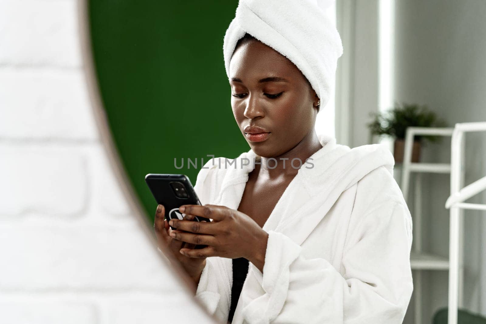 A woman enjoying her leisure time checks her smartphone while dressed in a soft bathrobe. The indoor space features a refreshing green backdrop and minimal decor.