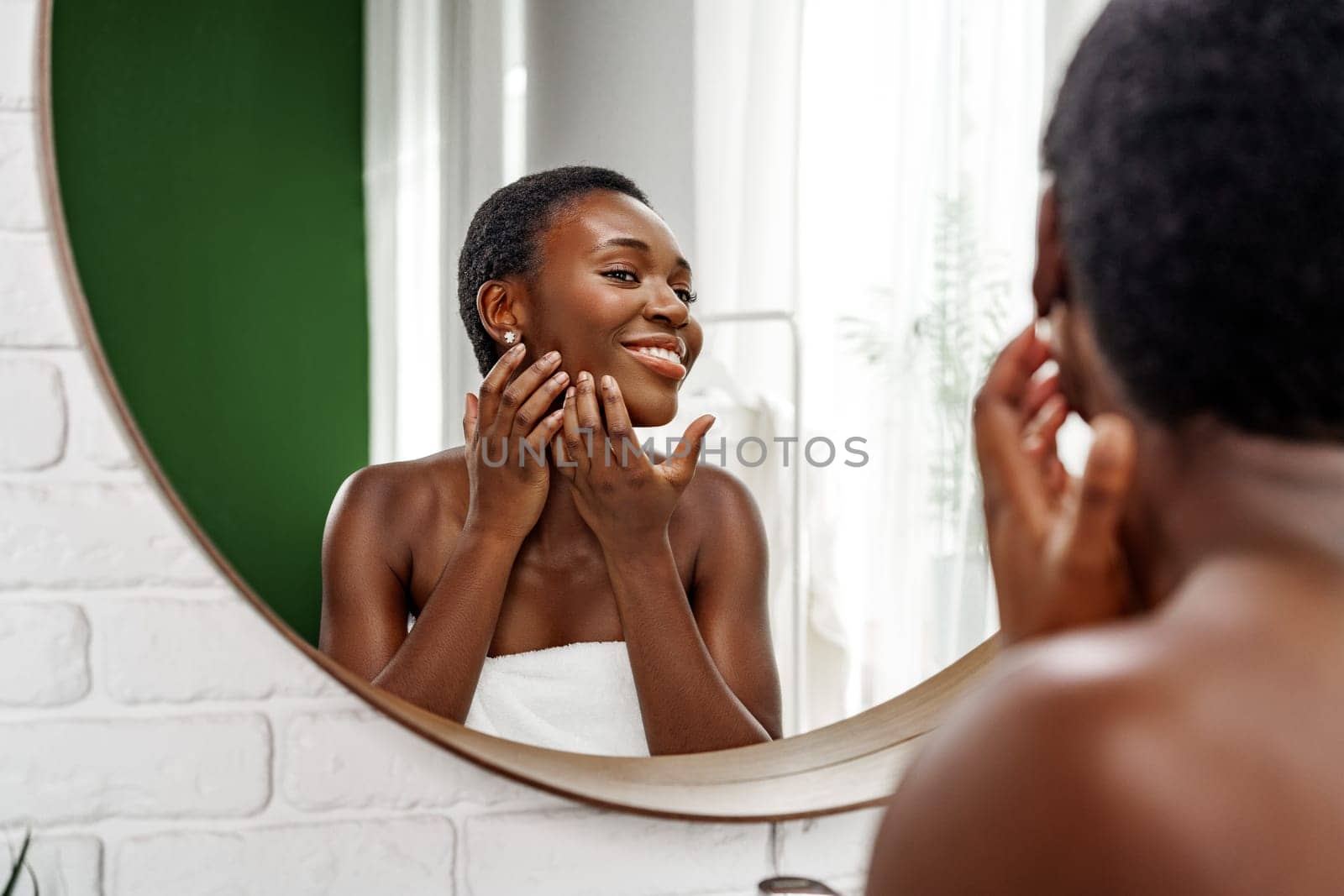 A woman with short hair smiles while applying skincare in front of a round mirror. The bright and clean bathroom features green accents and soft natural light, creating a serene atmosphere.