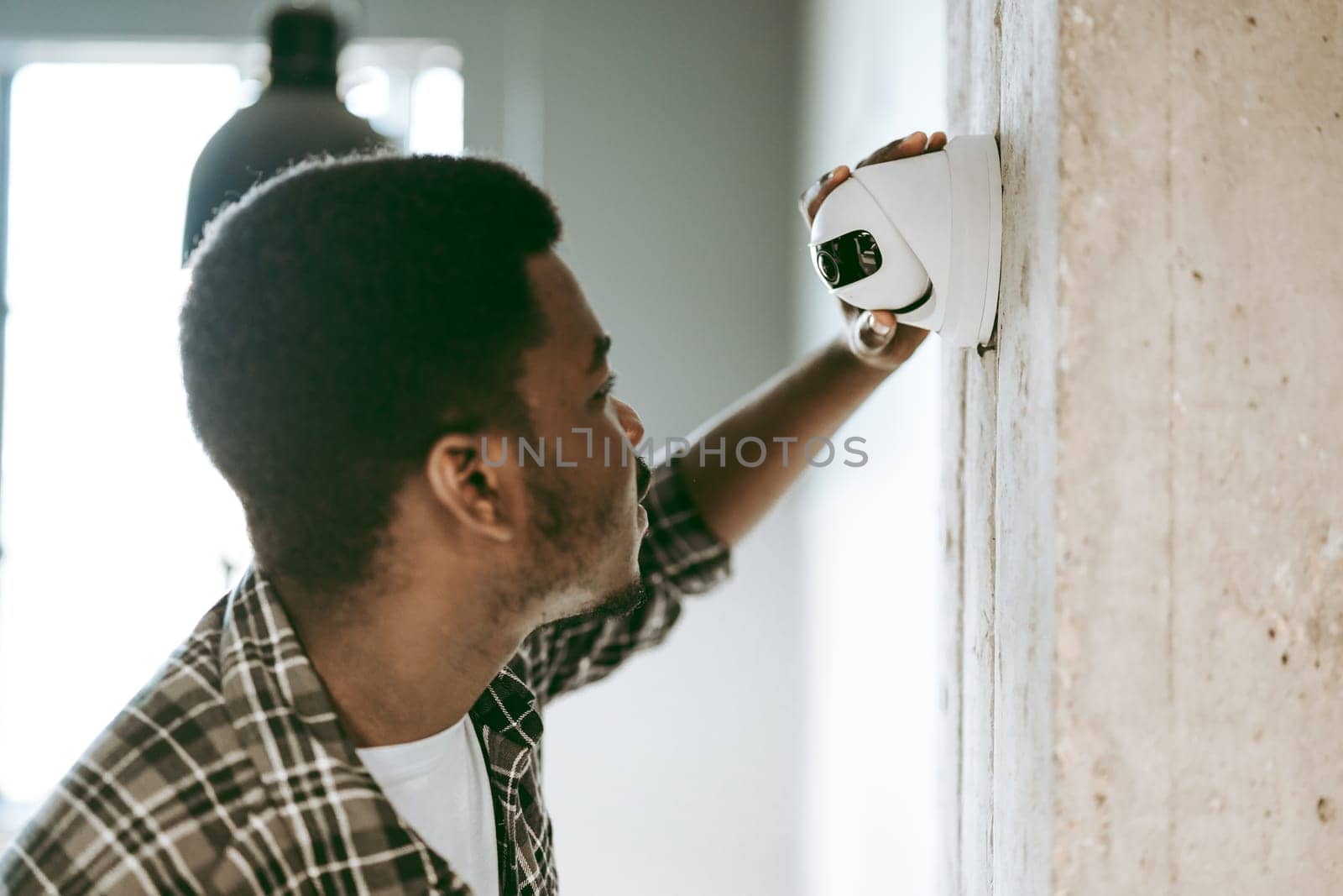 A young man is closely examining a newly installed security camera on a concrete wall in a bright, contemporary room.
