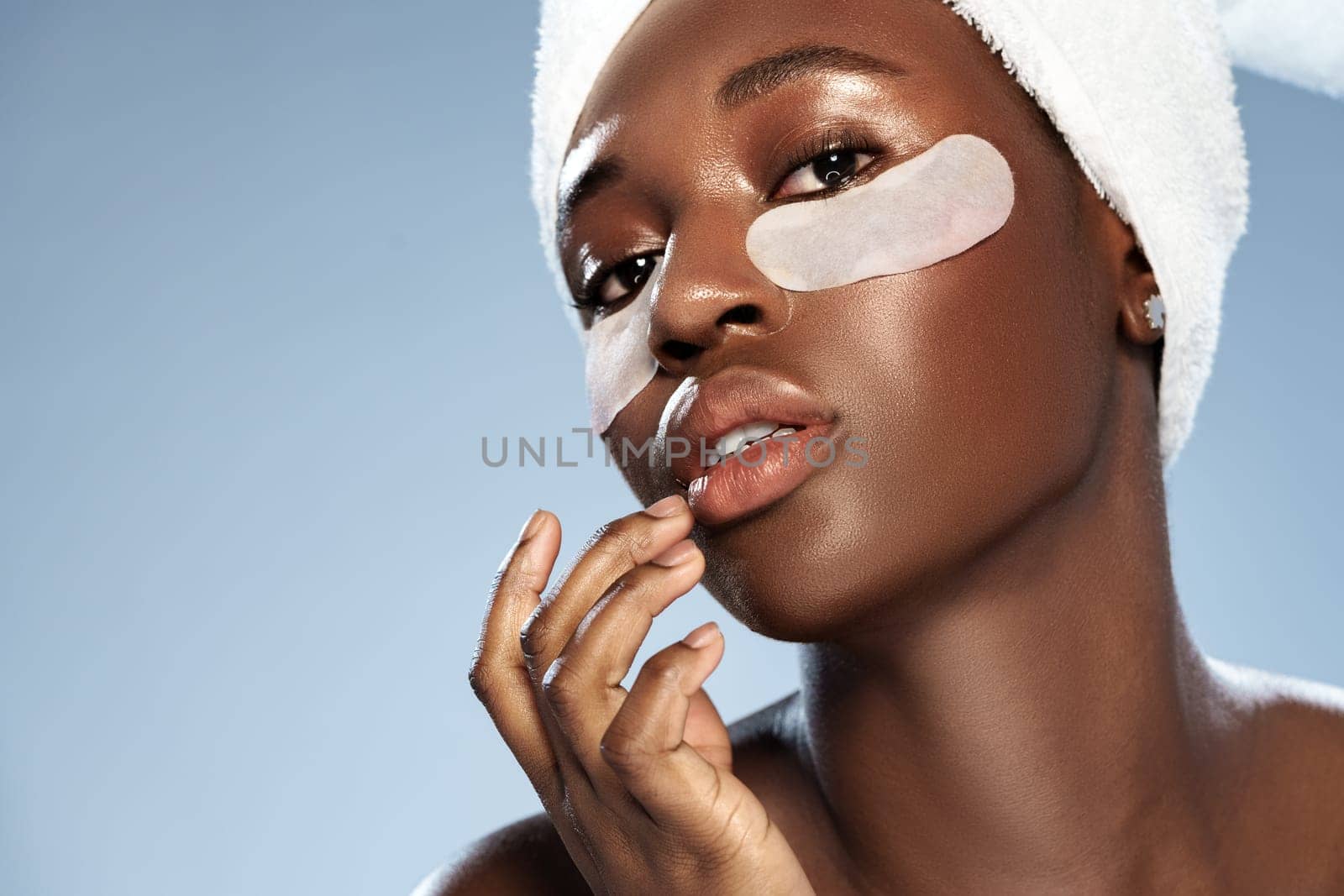 A woman with a towel wrapped around her head applies a finger to her lips. She wears eye patches and has a calm expression, embodying self-care and beauty rituals at home.