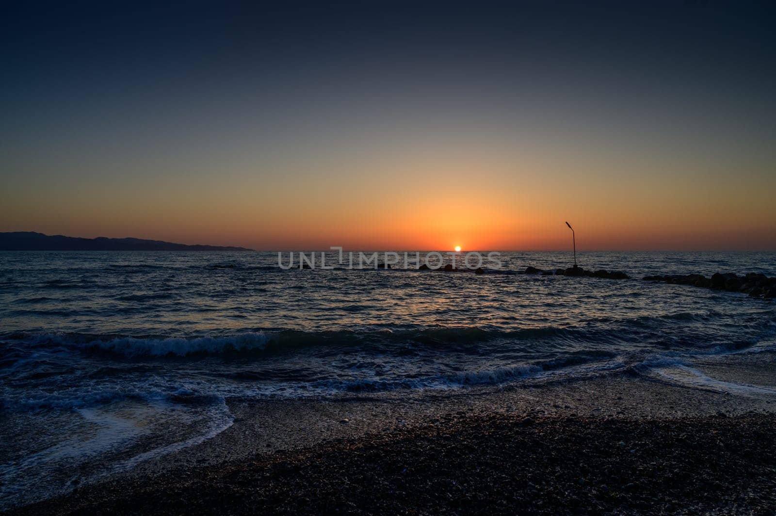 The sun sets into the calm waters of the Mediterranean Sea, casting warm reflec