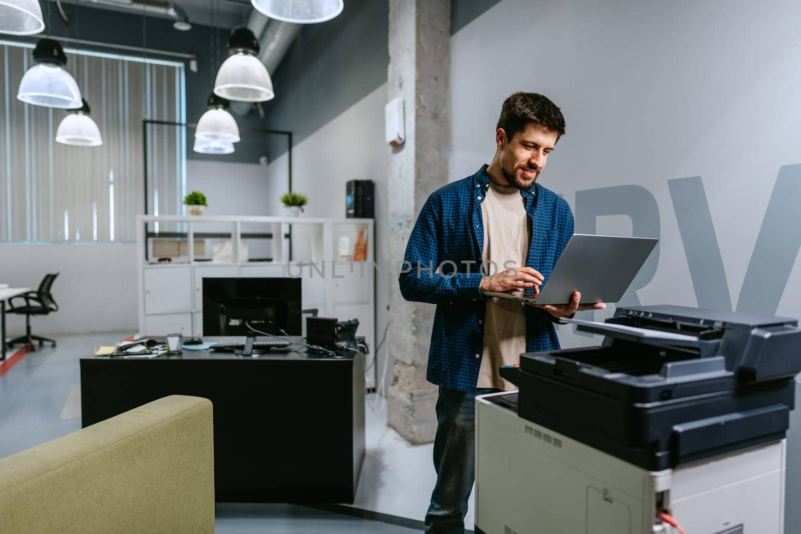 A young man stands by a printer in a modern office, working on his laptop. The space is bright and stylish, featuring contemporary decor and a relaxed atmosphere.