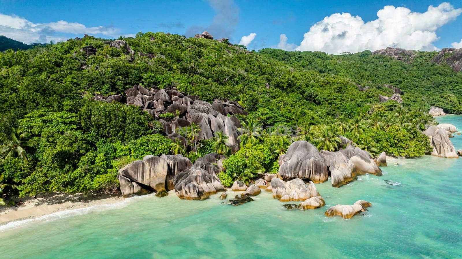 Turquoise water meets rocky cliffs and lush greenery in a panoramic coastal scene. La Digue, Seychelles.