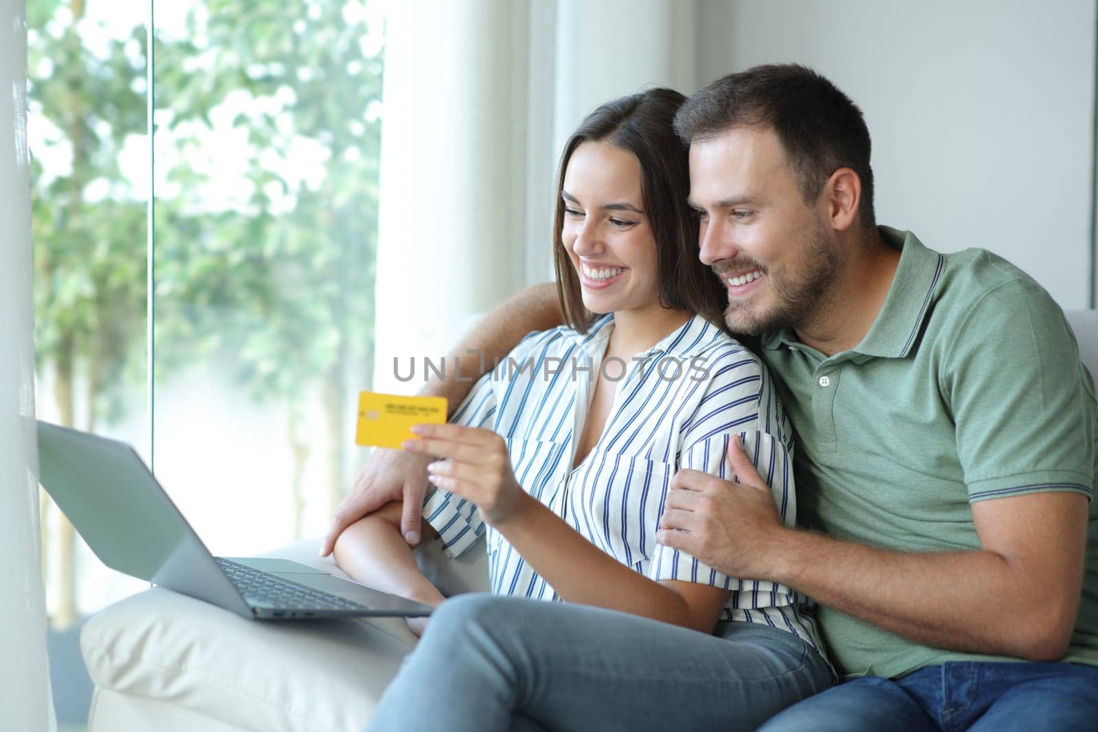 Happy couple buying online together with laptop and credit card sitting in a couch at home