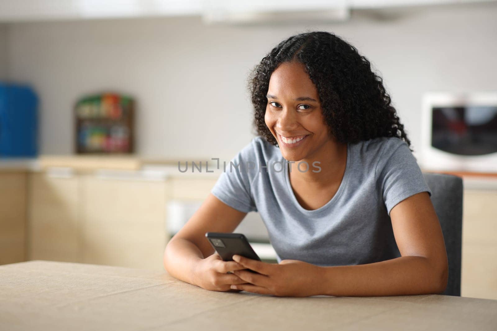 Happy black woman holding mobile phone looks at camera in the kitchen at home