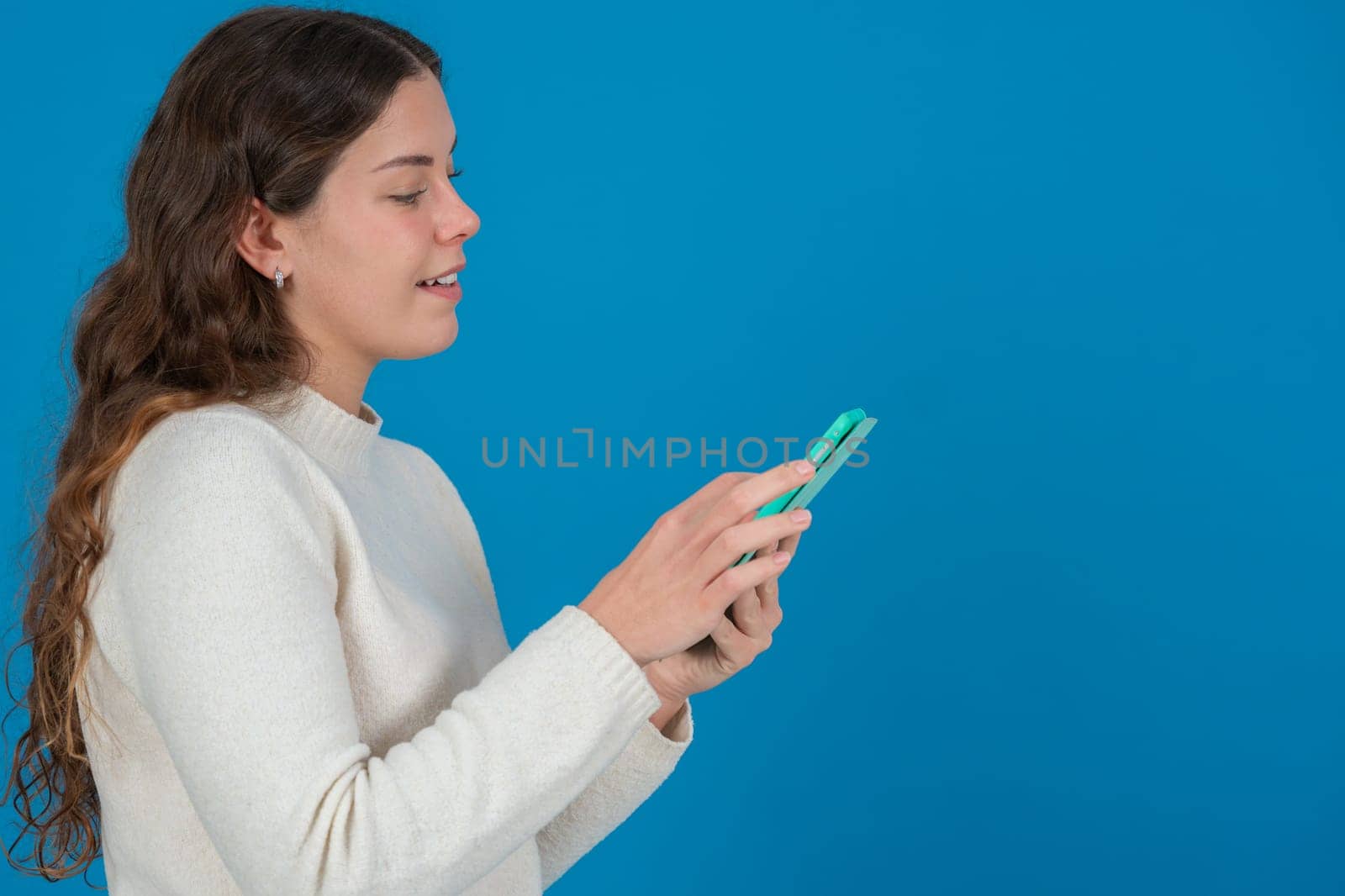 Side view of a smiling young woman in white sweater using a green tablet against a solid blue background with copy space, symbolizing digital communication, browsing, and technology interaction.