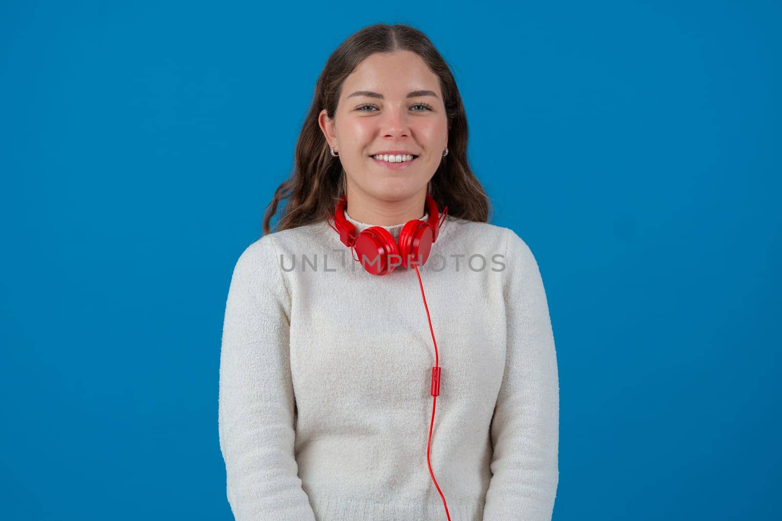 Individuality portrait Young woman wearing red headphones around her neck, smiling happily and looking relaxed. She appears joyful and confident. Solid blue background.